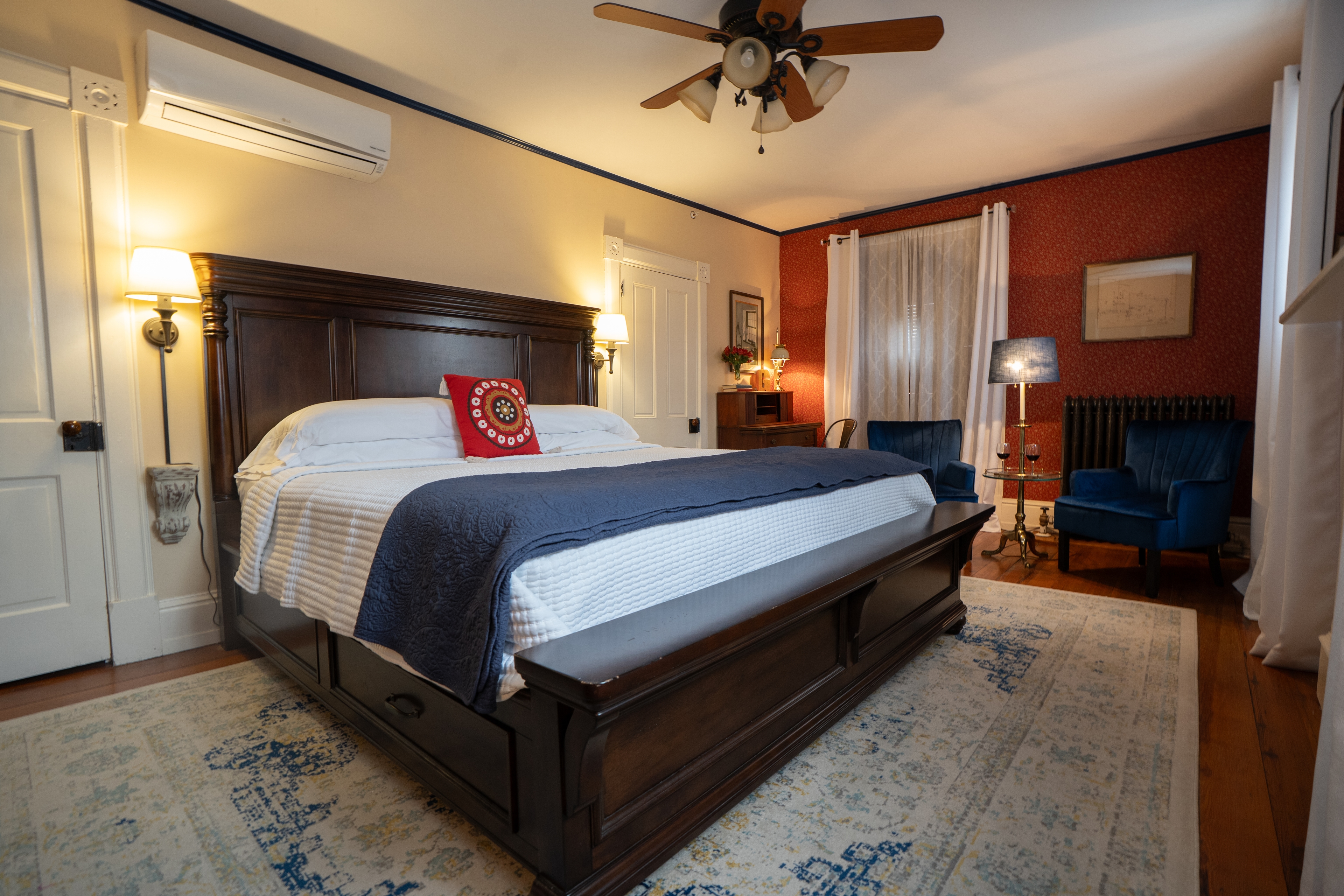 A king-sized bed with white linens and blue accents sits on a patterned rug in a historic bedroom.