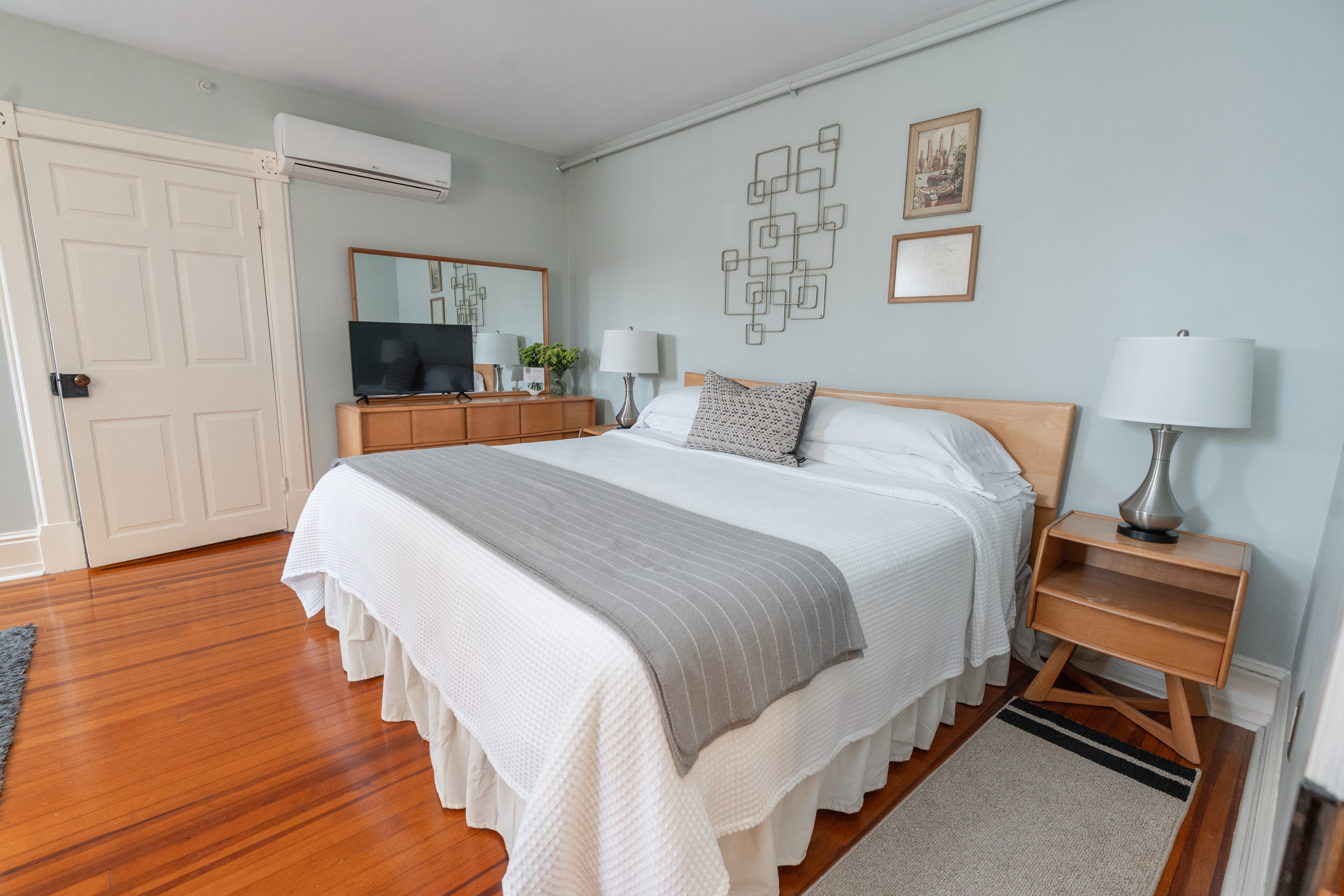 A mid-century modern guest room featuring a king bed with white linens and a brown plaid runner, light wood furniture, and soft blue walls.