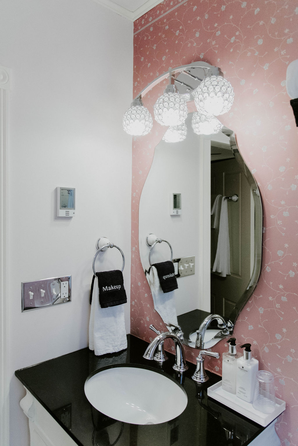 A modern bathroom vanity with a black countertop and white oval sink, featuring a unique scalloped mirror set against pink-and-white patterned wallpaper and illuminated by a multi-bulb globe light fixture.