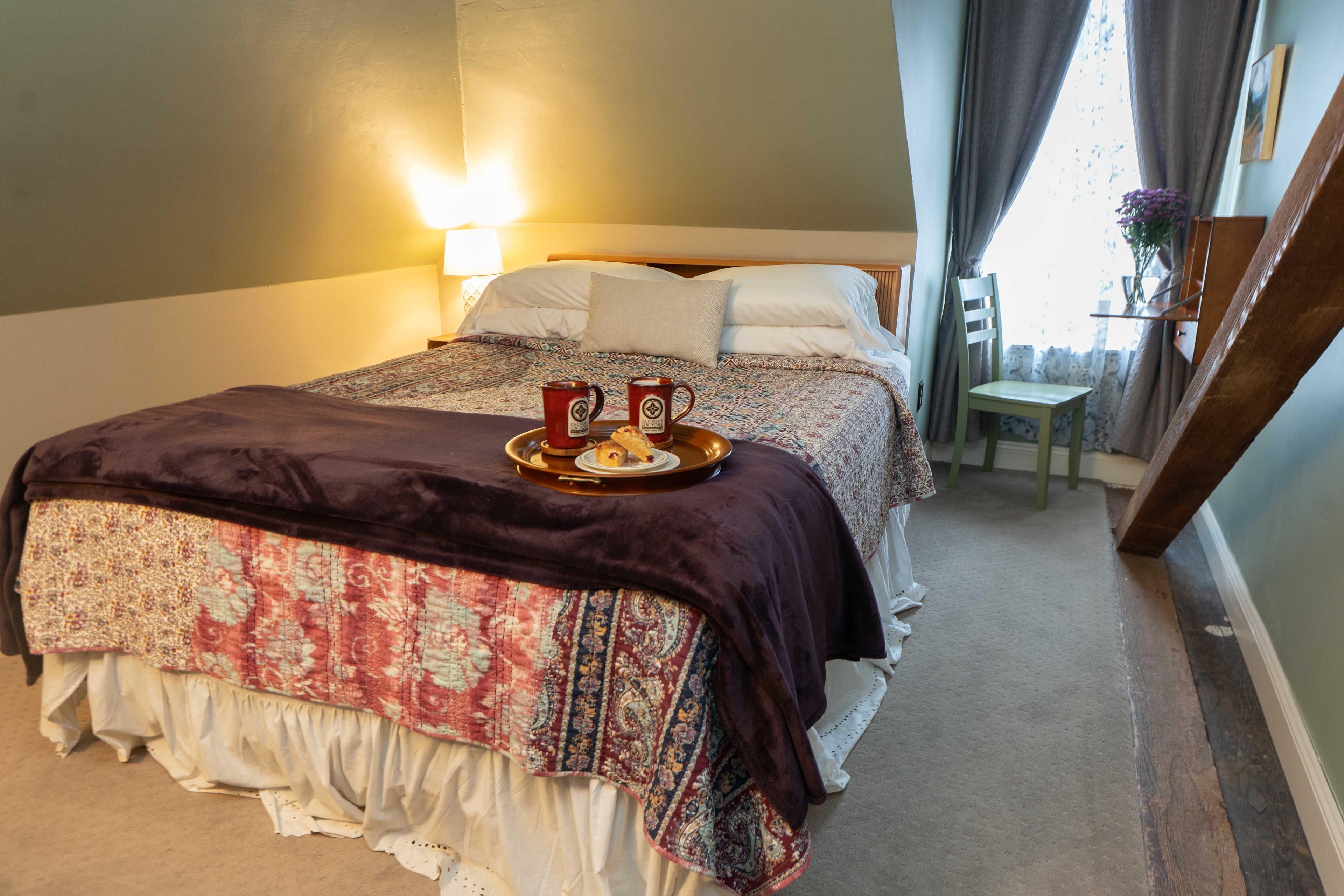 A queen bed with a red and white patterned quilt sits in a cozy attic guest room featuring sloped ceilings, a wooden ceiling beam, a vintage dresser, and a wall-mounted TV.