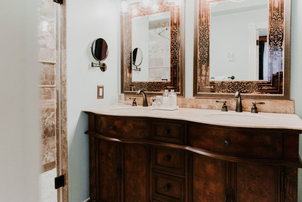 A double-sink vanity with dark wood cabinetry and ornate framed mirrors in a brightly lit luxury bathroom featuring a white countertop and oil-rubbed bronze faucets.