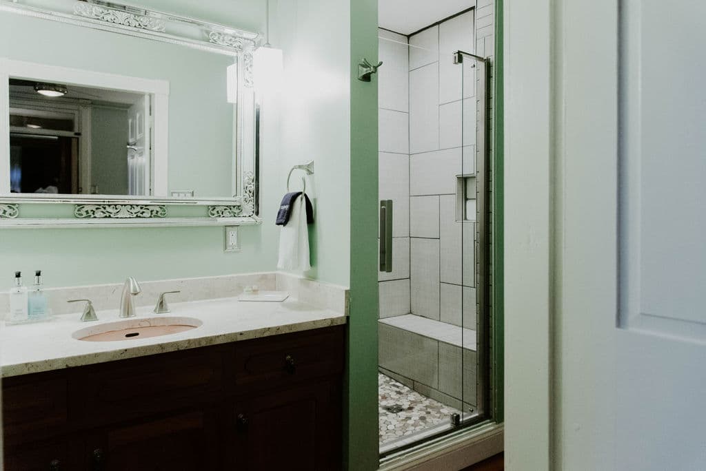 A brightly lit, modern luxury bathroom featuring a dark wood vanity with a white countertop and a glass-enclosed walk-in shower with light gray tiling and a built-in bench.