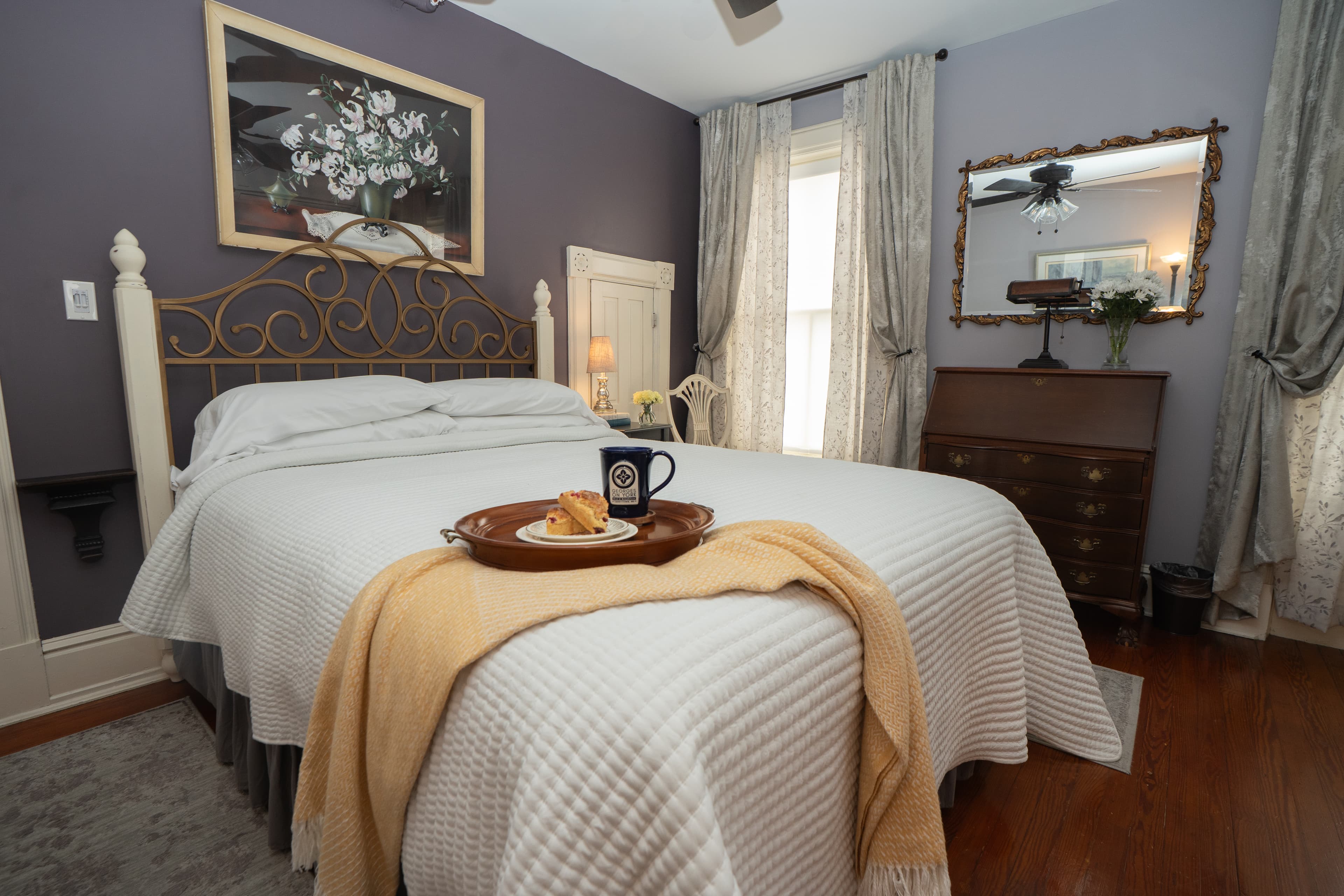 Wilt Room queen size bed with a gold ornate metal headboard, white bedding, and a decorative floral painting hanging on a dark purple wall above.