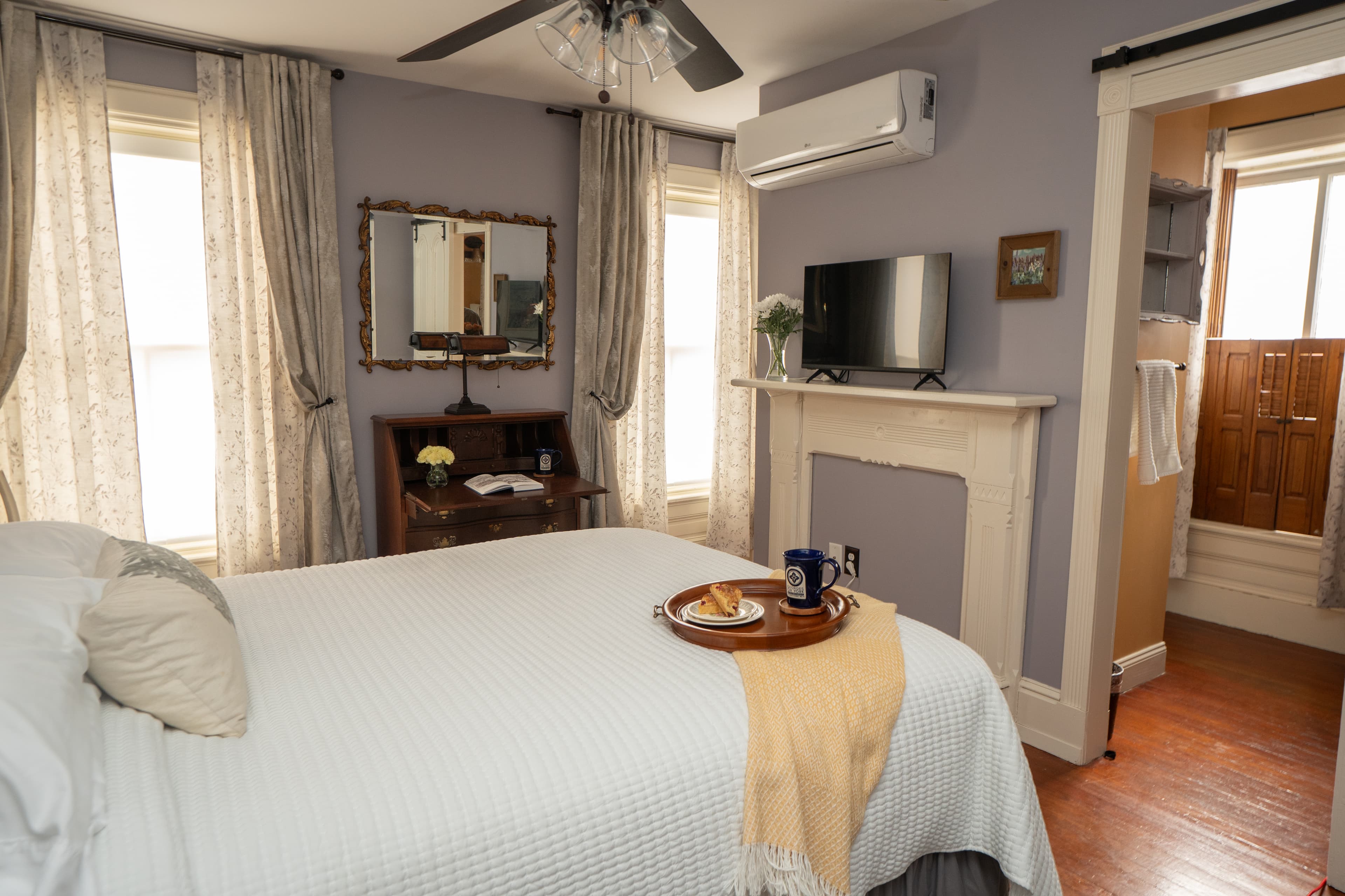 A cozy bedroom featuring a queen bed with a decorative gold scrollwork headboard, white linens, and a yellow throw blanket, with two windows and a ceiling fan.