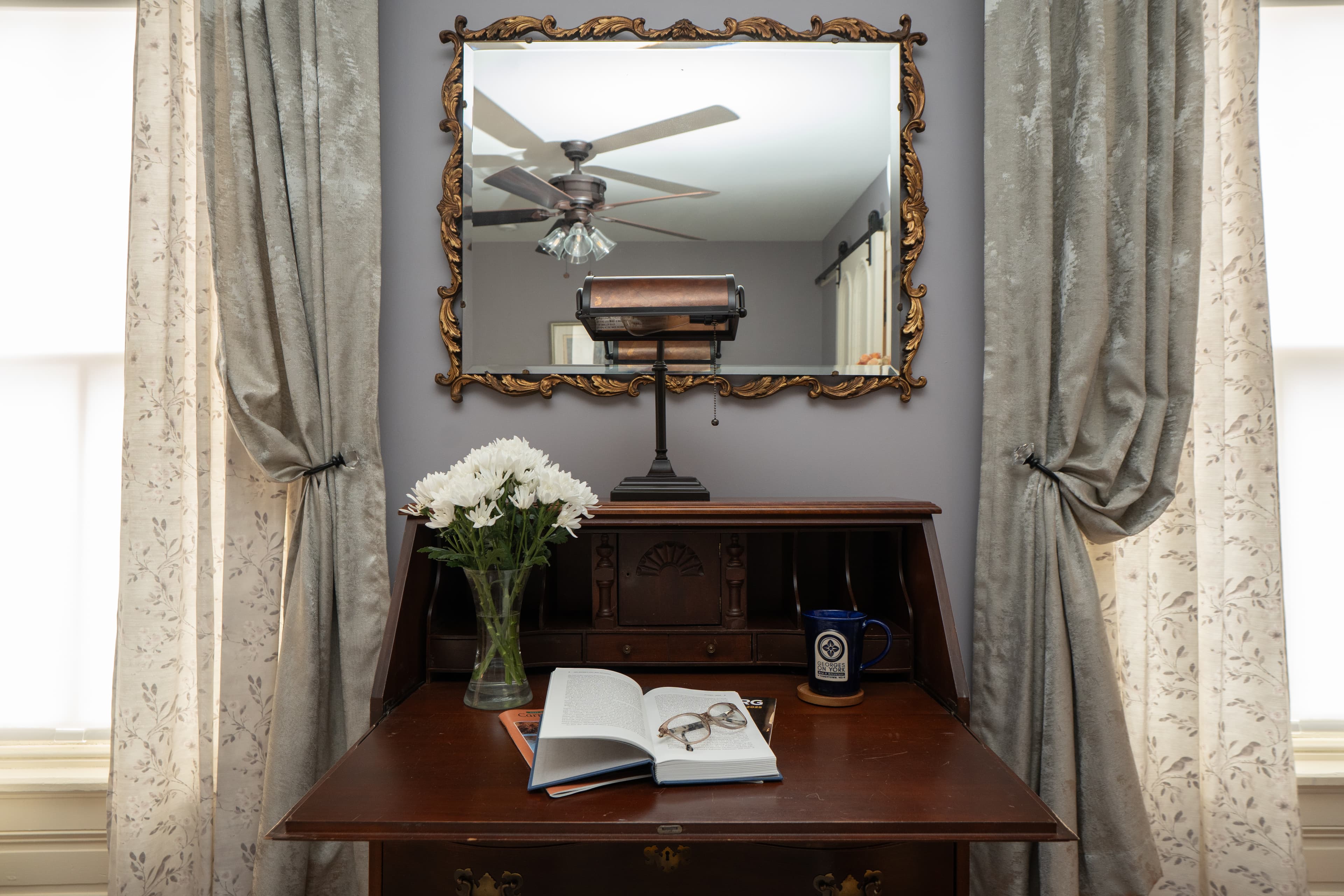 A vintage dark wood secretary desk with a brass lamp sits in a room with light purple walls, featuring a white fireplace mantel and an air conditioning unit in the background.