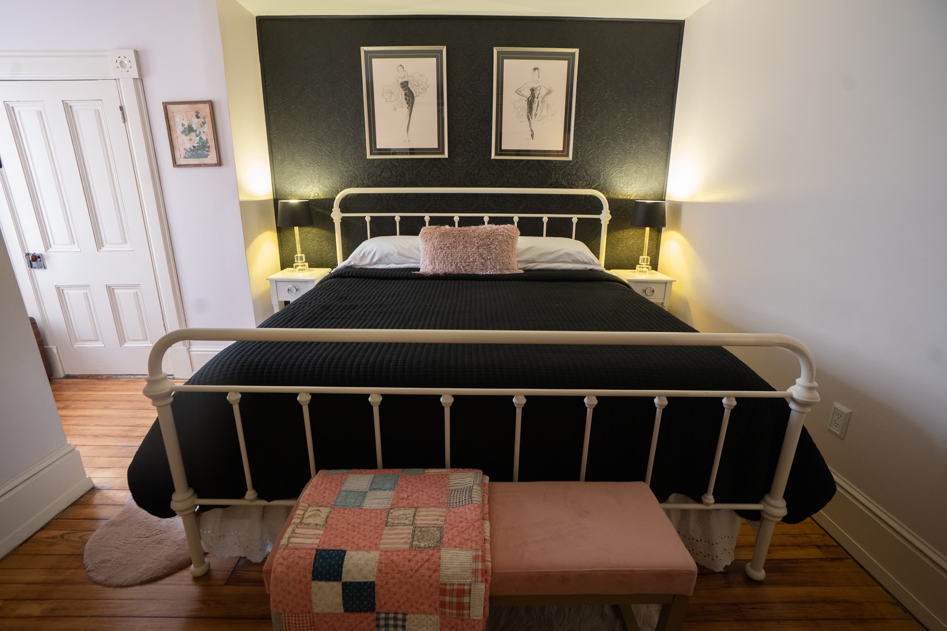 A queen-sized white metal bed frame with black linens is centered against a black accent wall featuring two framed minimalist art prints.