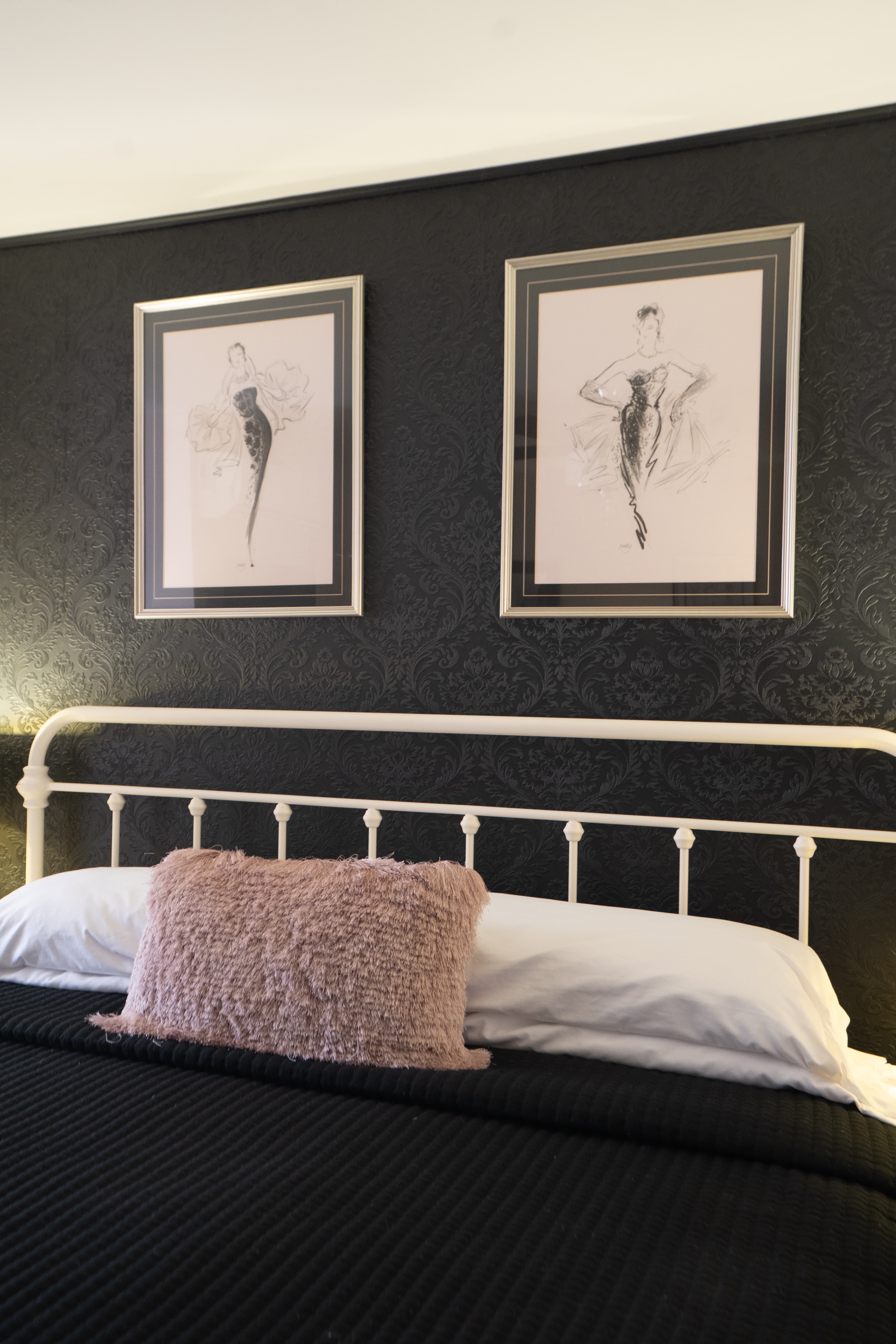 A close-up of a white metal headboard and bed with black and white linens, featuring a textured pink decorative pillow in the foreground.