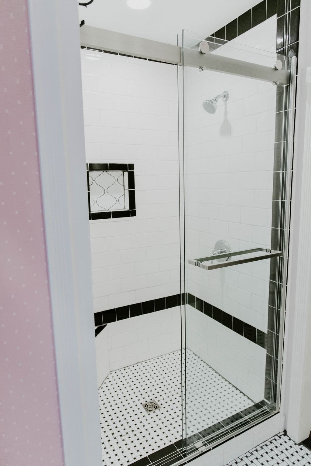 A glass-enclosed walk-in shower with white wall tiles, black accent border tiles, and a black-and-white penny tile floor.