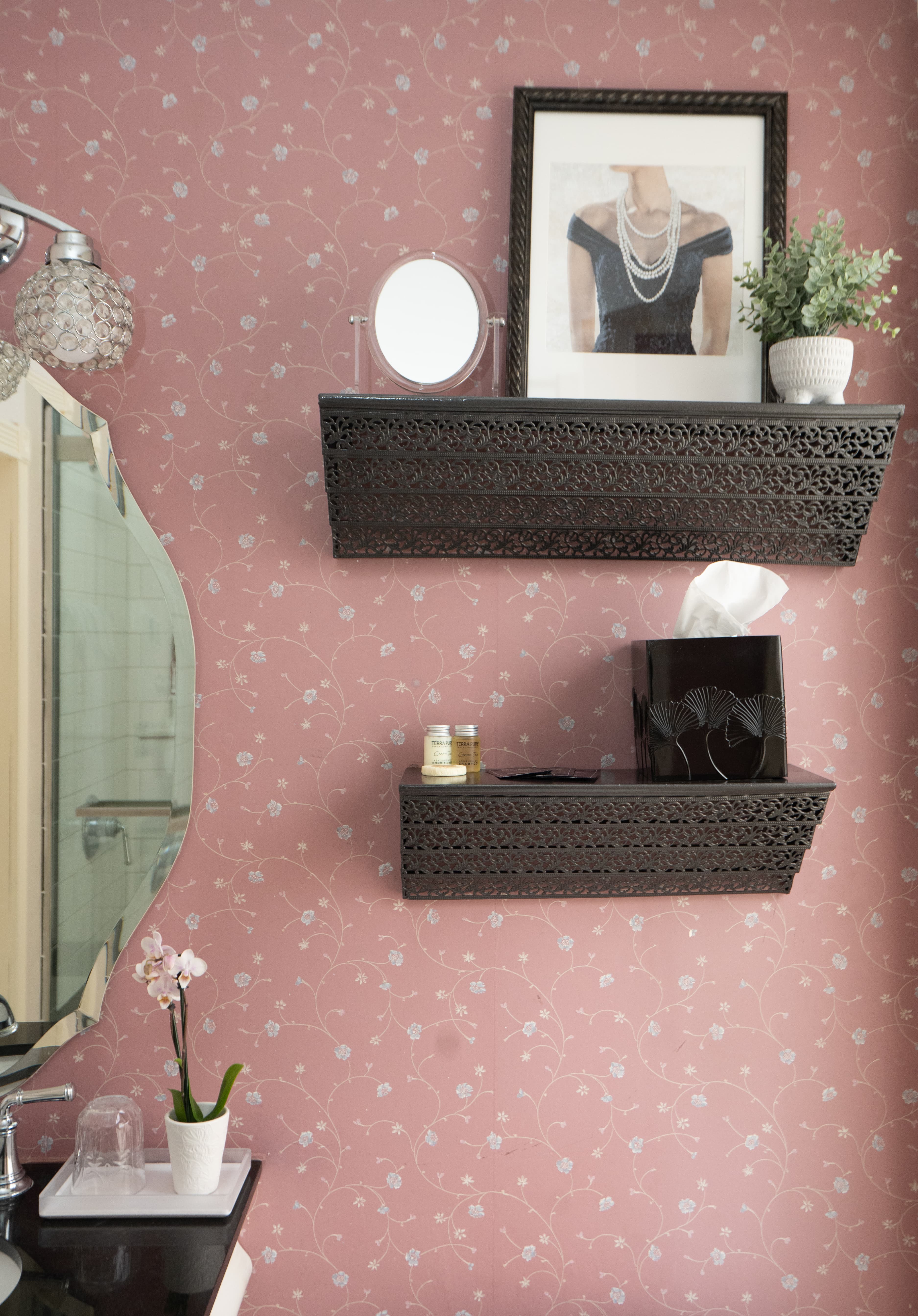 Two decorative, black wall shelves with an intricate cut-out pattern. The lower shelf holds a small round mirror and a folded white towel against pink-and-white patterned wallpaper.