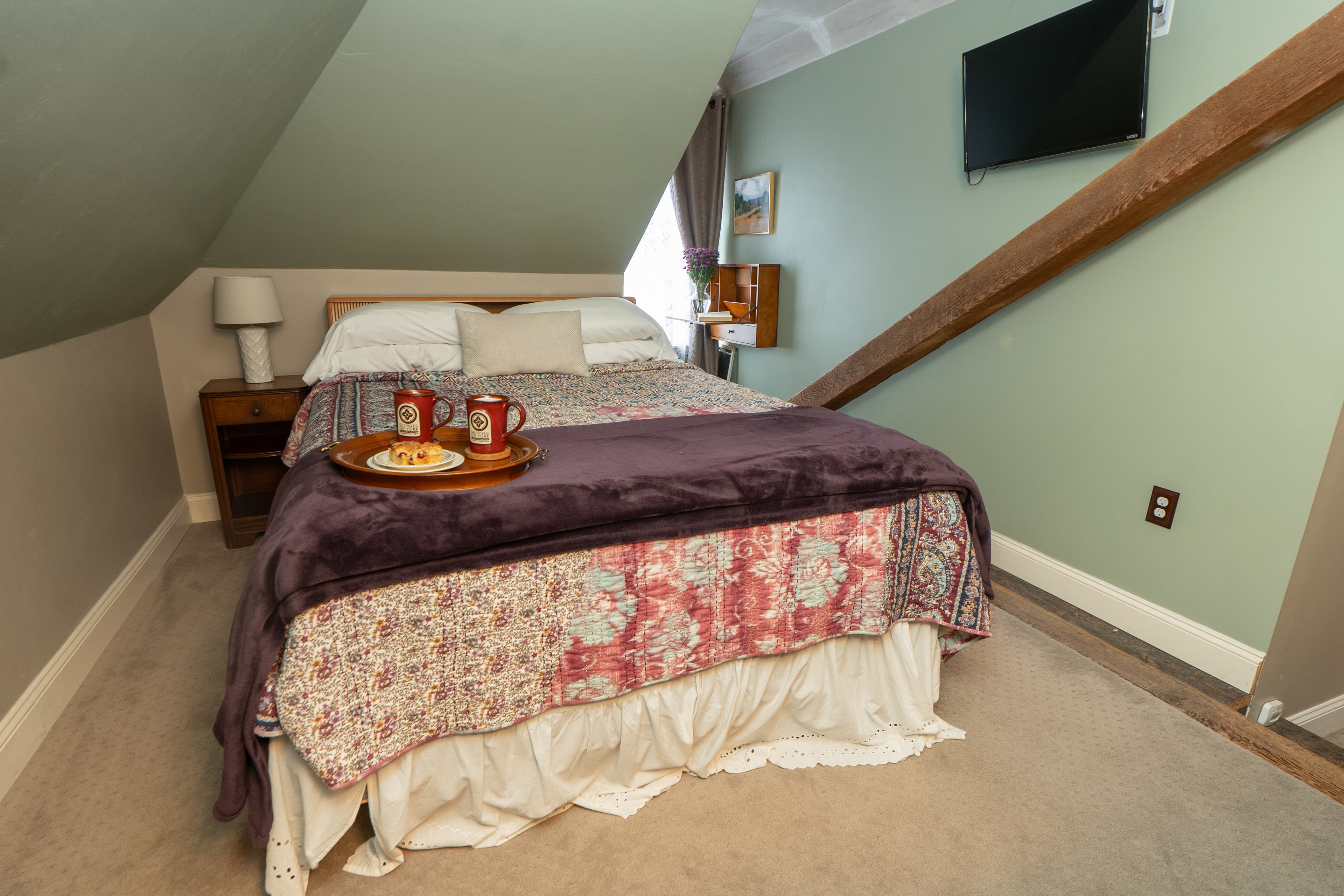 A cozy attic bedroom with sloped ceilings featuring a queen bed covered in a red, grey, and white patterned quilt, alongside a light wood dresser.