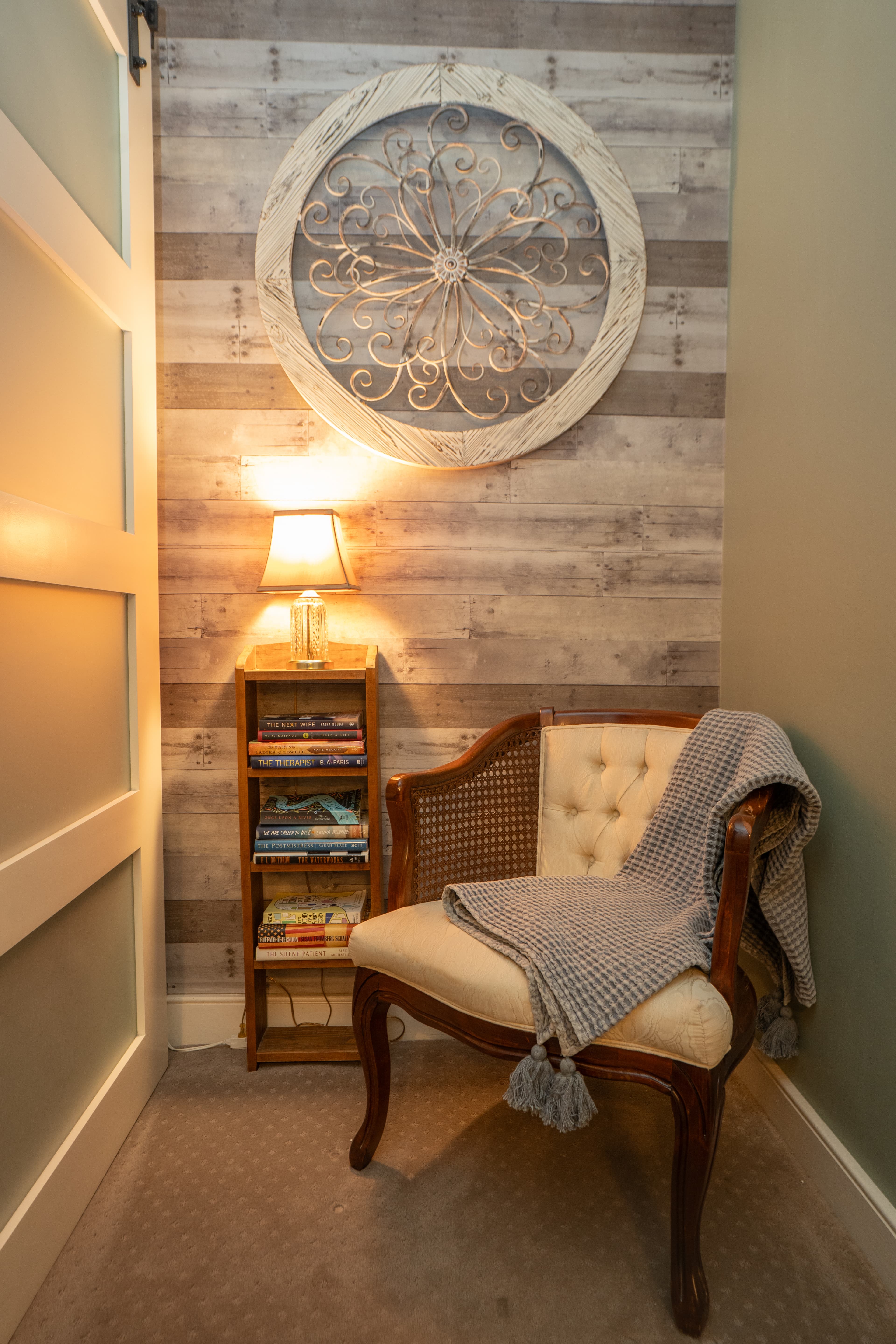 A white upholstered chair sits in a corner with a rustic wood-plank accent wall, featuring a large circular carved wooden wall hanging and a small wooden shelf with books and a lamp.
