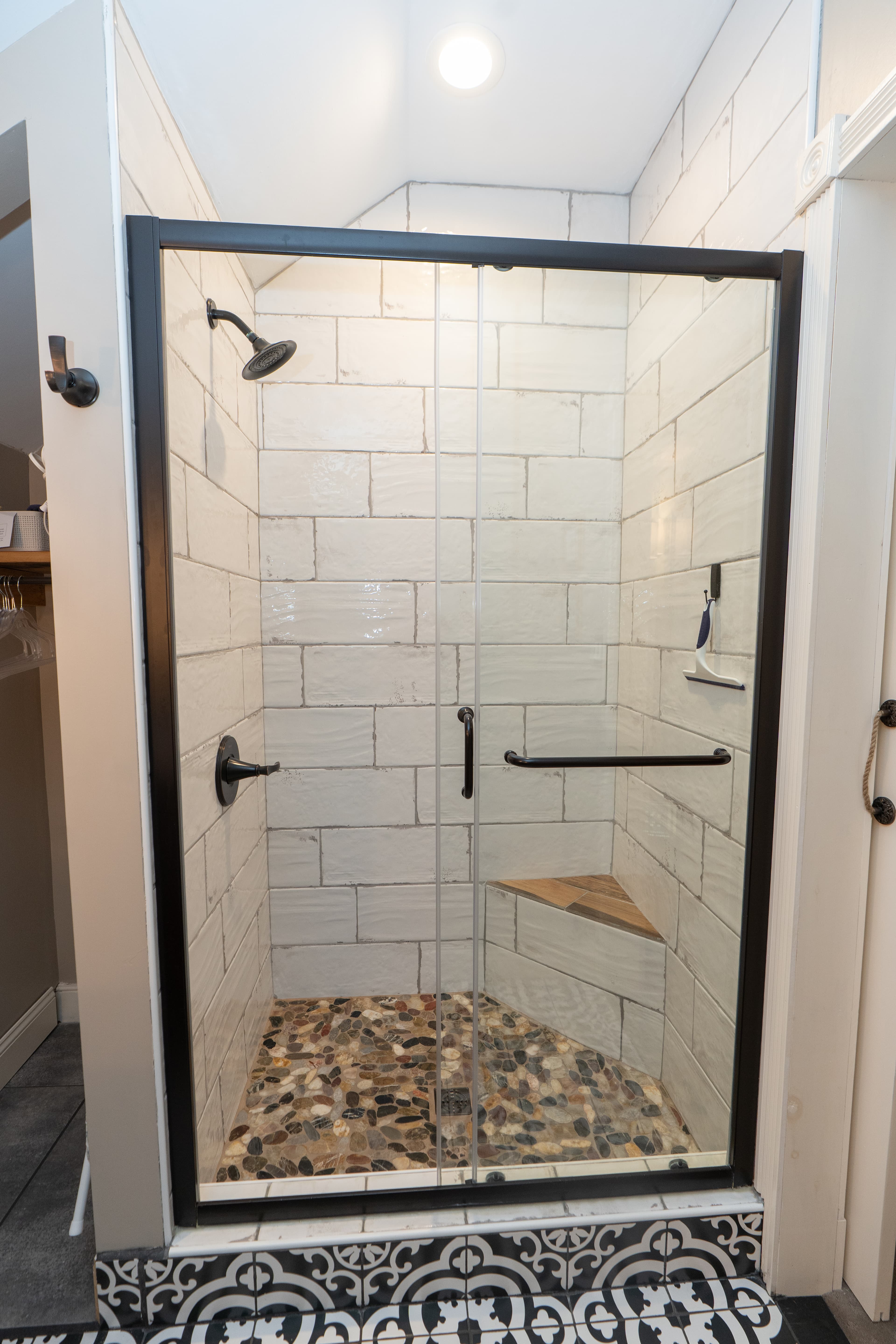 A glass-enclosed walk-in shower featuring classic white subway wall tiles, a pebble-stone floor, and black hardware, set against a black-and-white patterned tile bathroom floor.