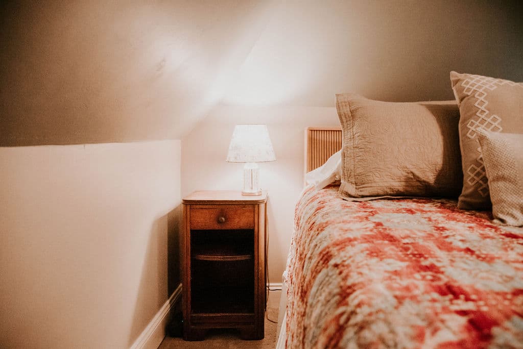 A close-up of a small, vintage-style wooden bedside table with a lit lamp, positioned next to a bed with a red-and-white patterned quilt in a cozy attic room.