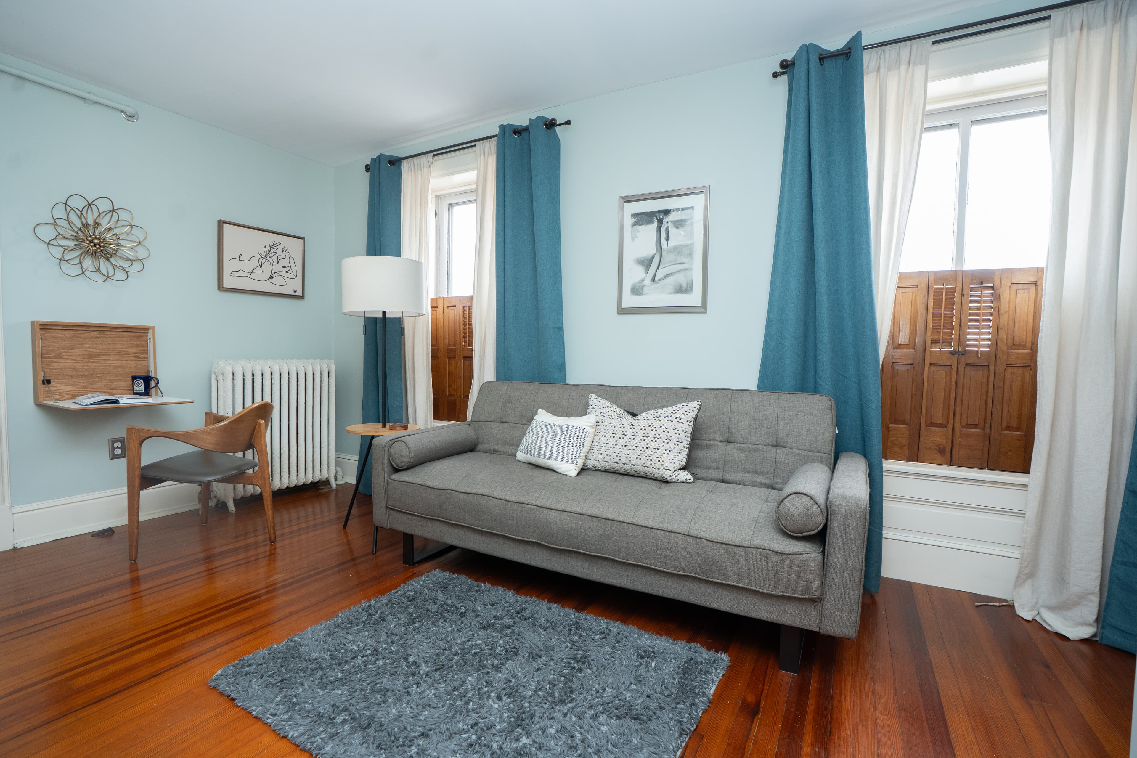 A seating area in a mid-century modern guest room featuring a grey sofa, a wooden accent chair, and a gold floor lamp set against blue curtains and hardwood floors.