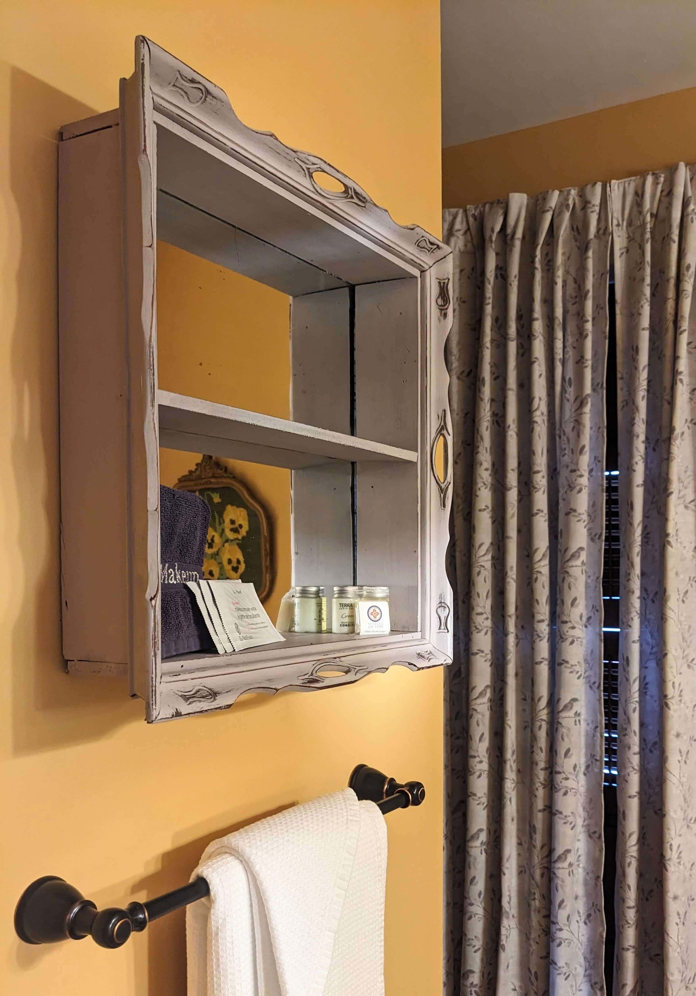 A vintage wall shelf with toiletries and towels, set against a yellow wall and floral curtains.