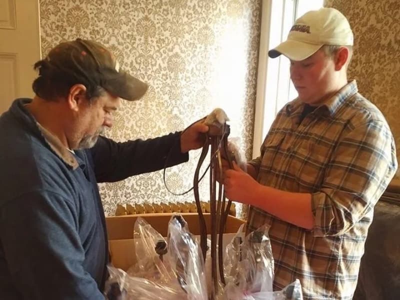Two men are sorting through horse tack in a room with patterned wallpaper.