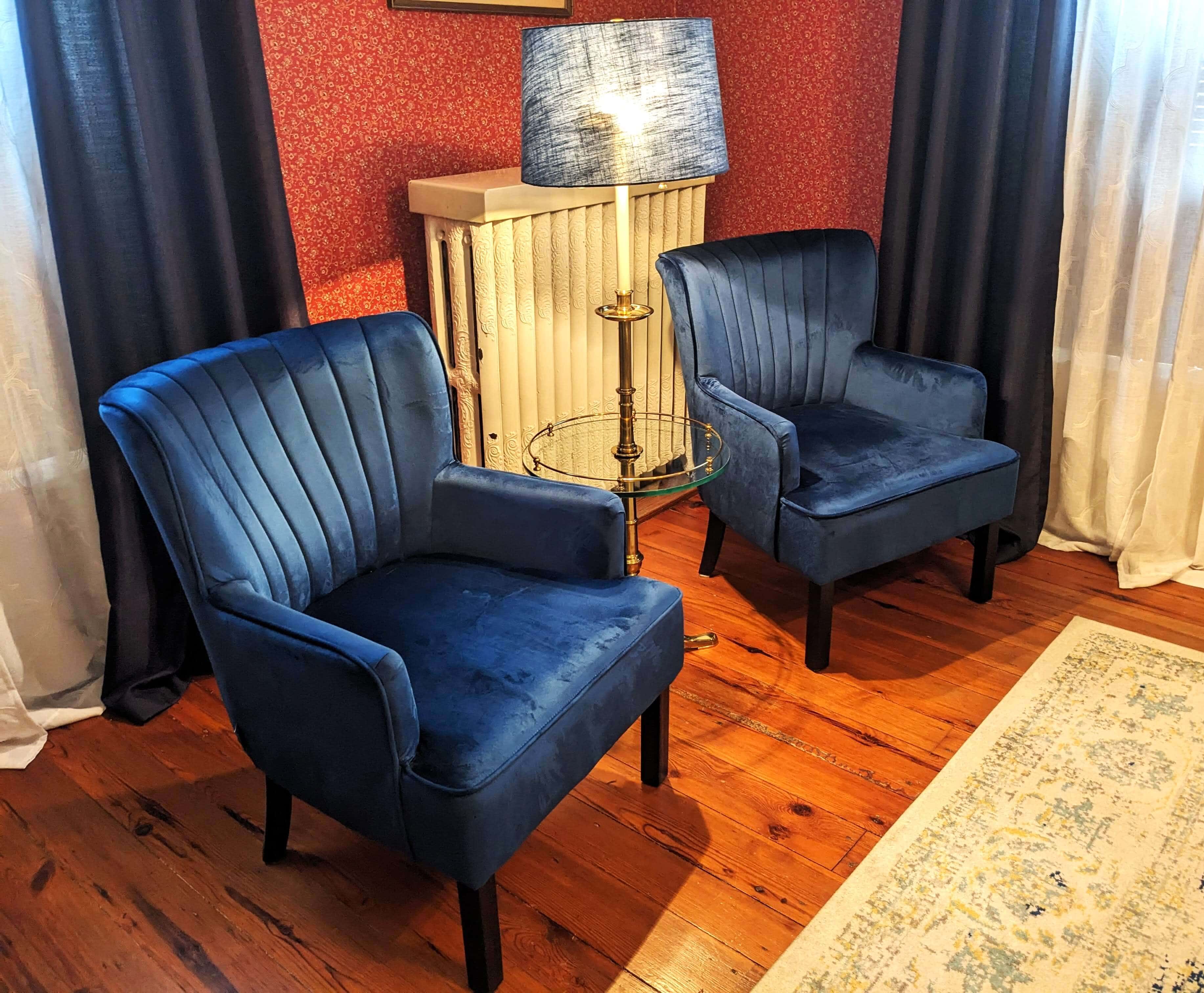 Two blue velvet chairs sit beside a small glass table and a lamp in a cozy room with patterned walls.