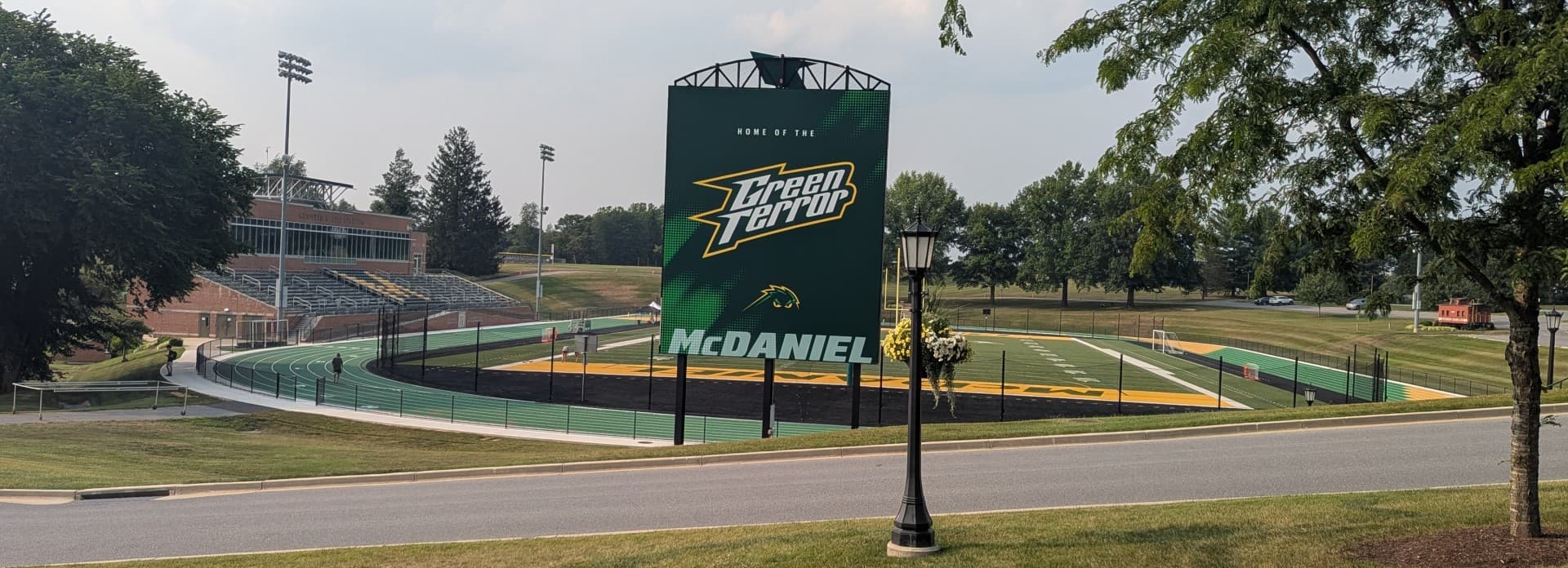 A large digital scoreboard displaying "Green Terror" and "McDaniel" at a sports field surrounded by trees.