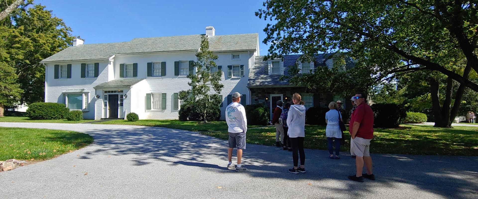 A group of people stands in front of a large, two-story white house on a sunny day.