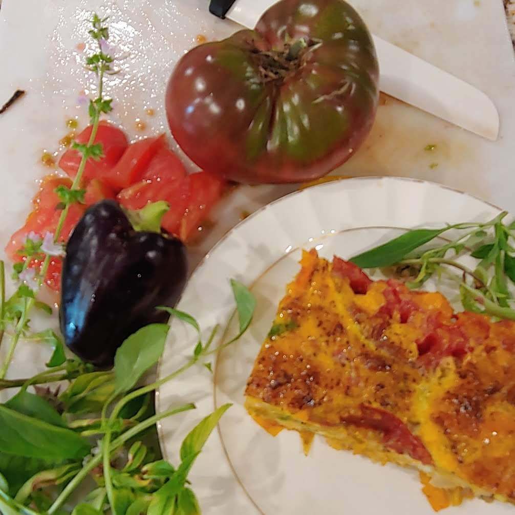 A slice of vegetable casserole is served on a plate alongside fresh tomatoes, a bell pepper, and herbs on a cutting board.