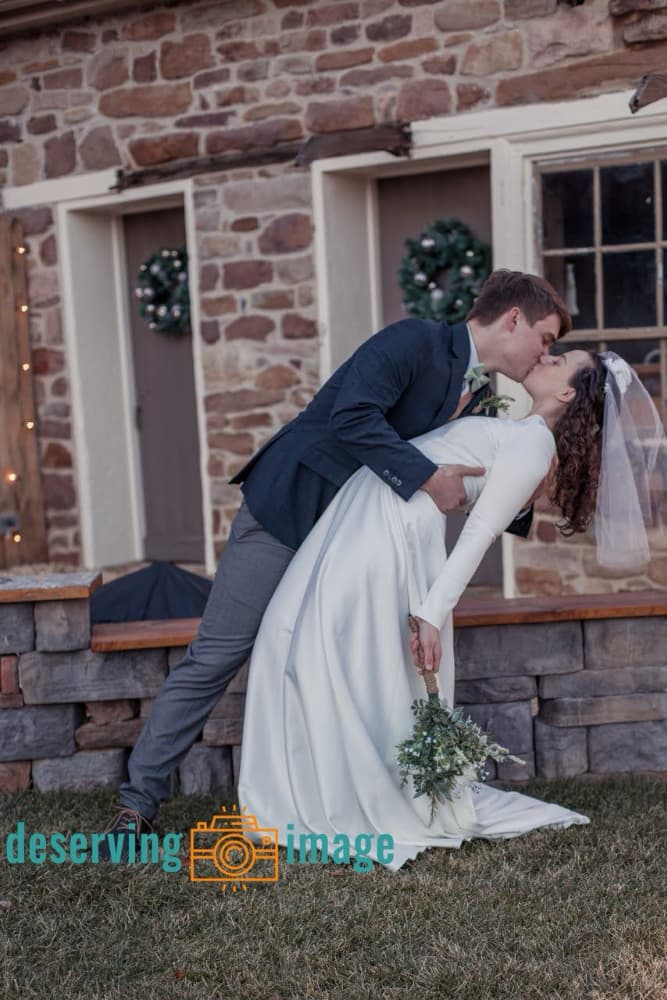 A newlywed couple shares a romantic kiss while posing outdoors in front of a rustic stone building.