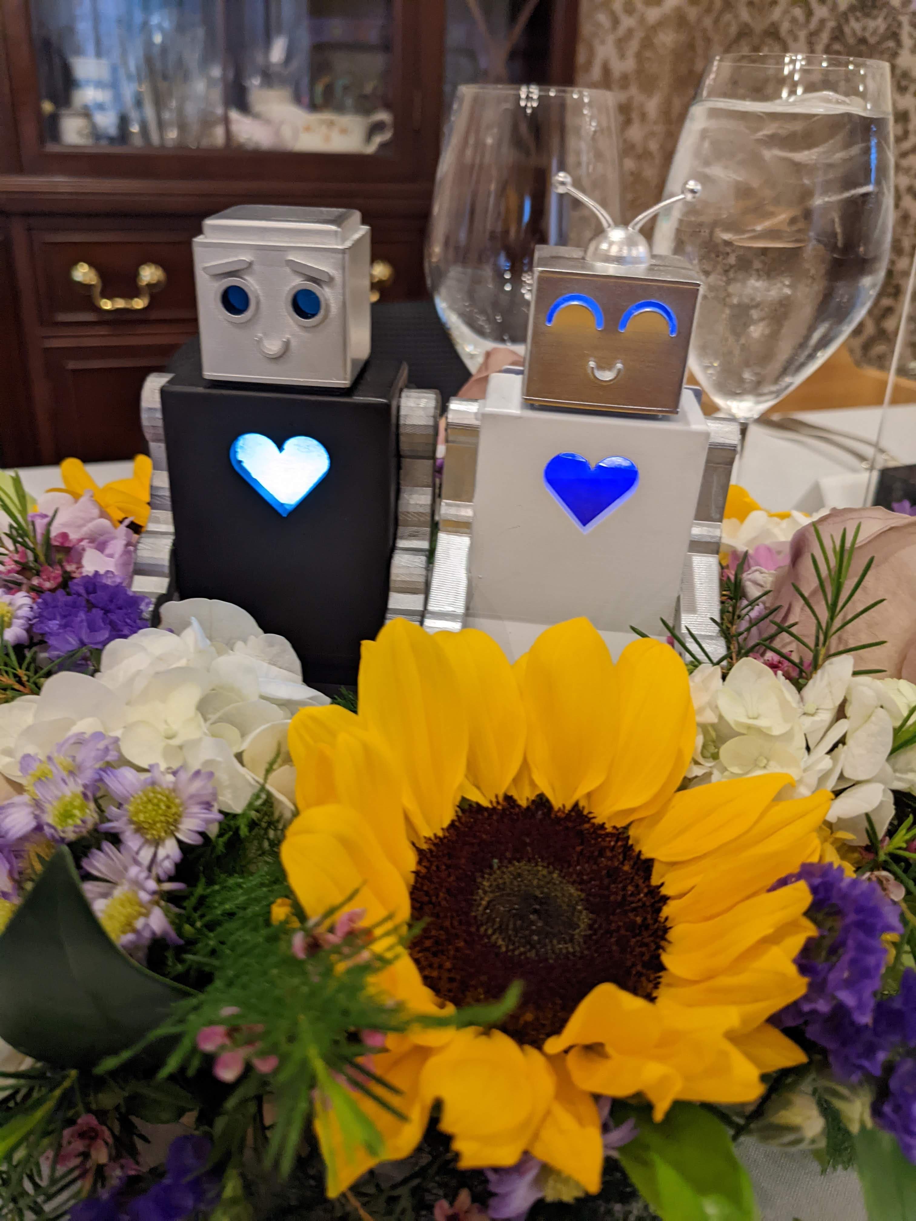 Two cute robots with heart-shaped lights sit among colorful flowers and a sunflower, flanked by glasses of water.