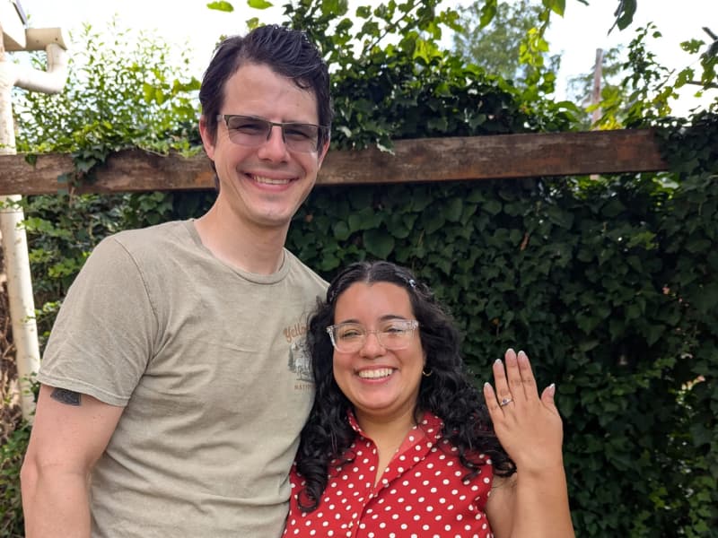 A smiling couple stands outdoors, with one showing off an engagement ring against a background of greenery. A smiling couple stands outdoors, with one showing off an engagement ring against a background of greenery.