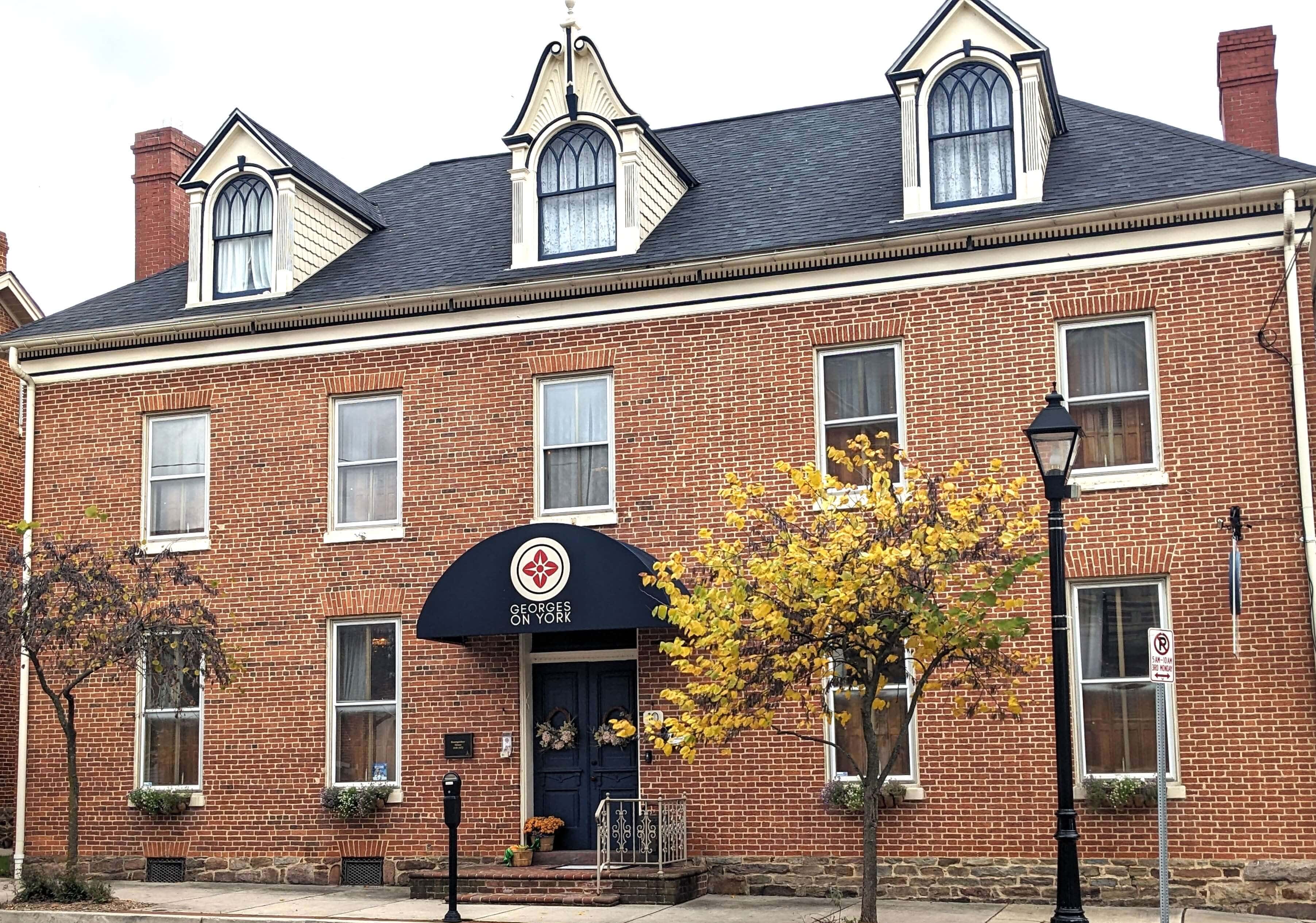 A brick building with a gabled roof, large windows, and a decorative entrance beneath an awning, surrounded by trees and a lamppost.