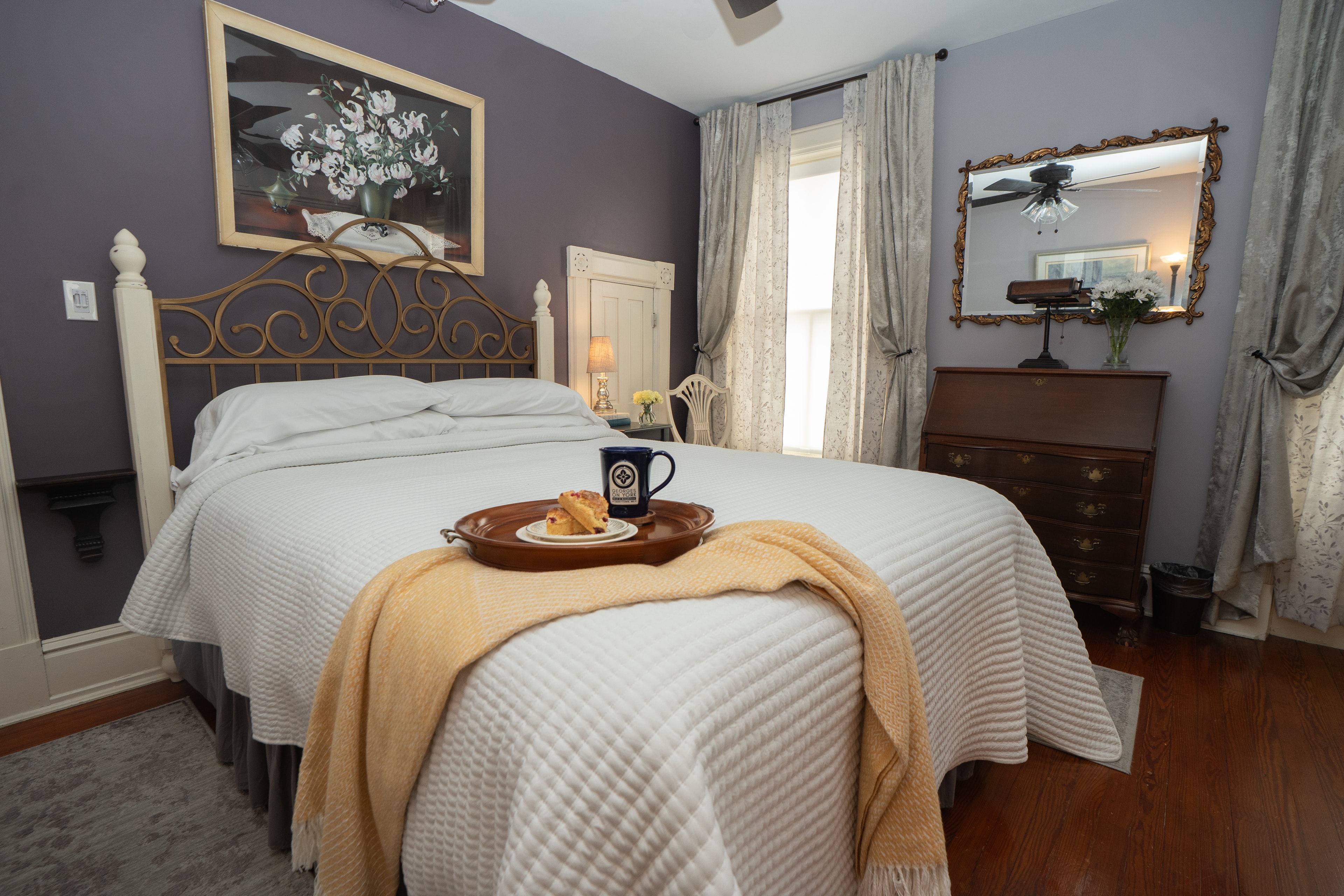 Wilt Room queen size bed with a gold ornate metal headboard, white bedding, and a decorative floral painting hanging on a dark purple wall above.