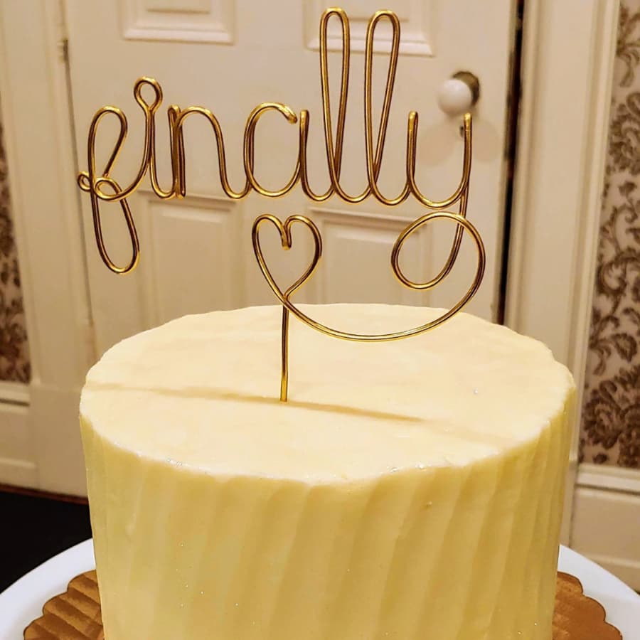 A round cake topped with a gold wire sign that reads "finally" with a heart.