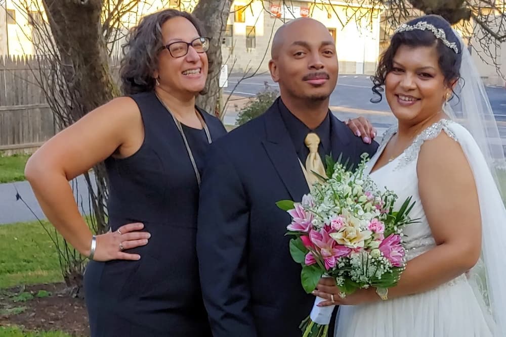A smiling bride holds a bouquet, standing with a groom and a woman in a black dress.