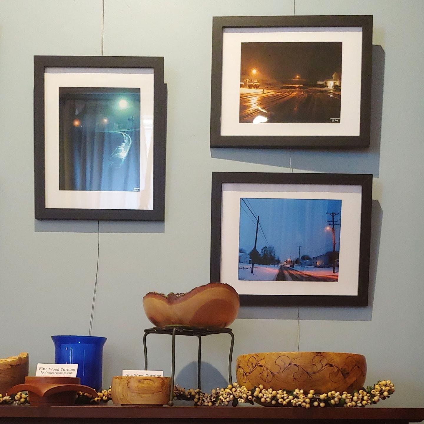 A display showcases three framed photographs on a blue wall, accompanied by decorative wooden bowls and a blue vase.