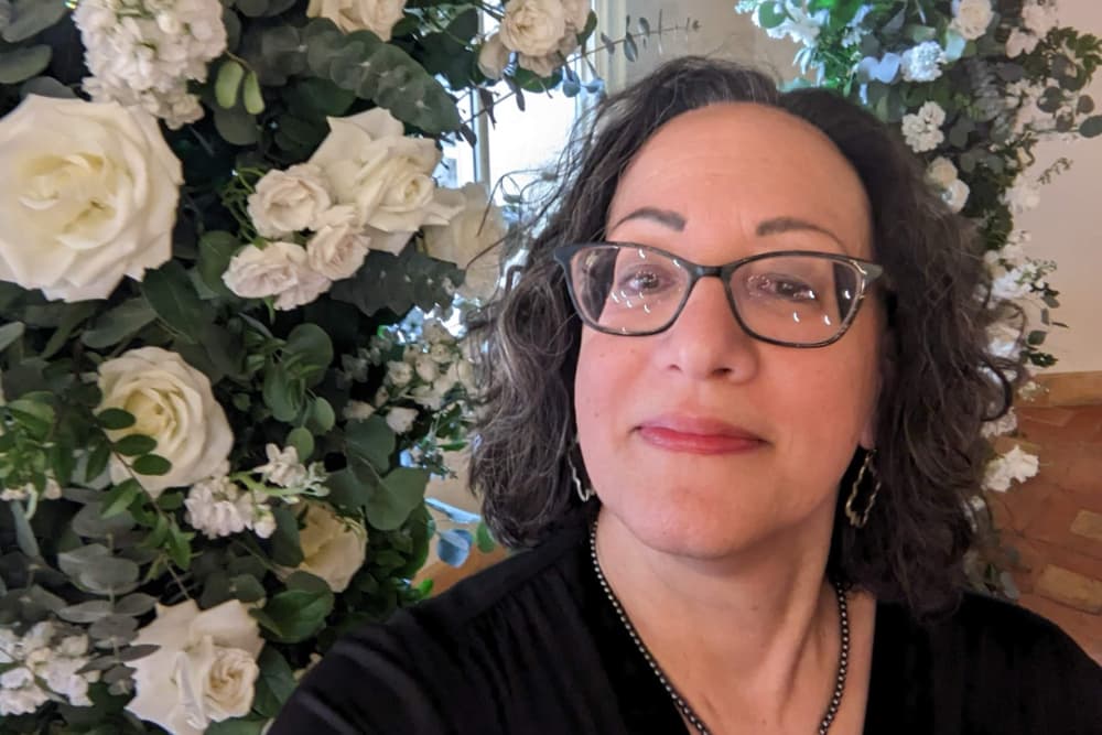 A woman with curly hair and glasses poses in front of a backdrop of white flowers.