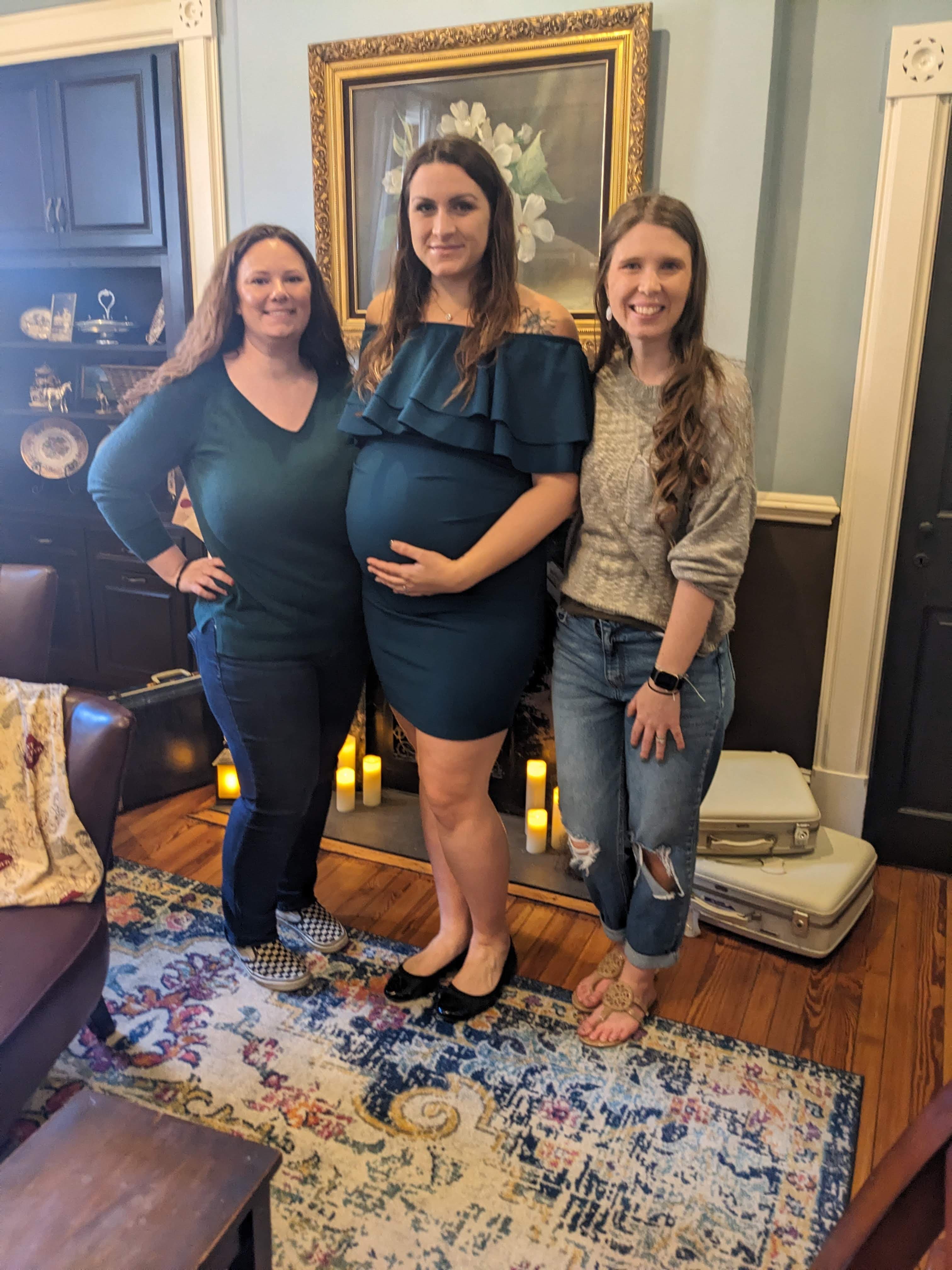 Three women pose together indoors, with one woman prominently displaying her pregnancy.