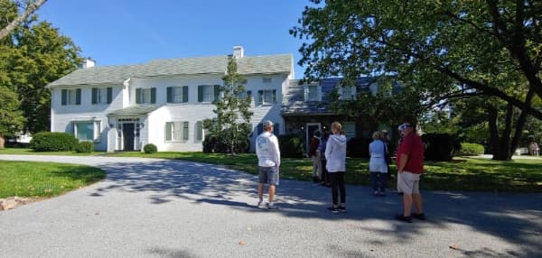 A group of people stands outside a large white house on a sunny day.