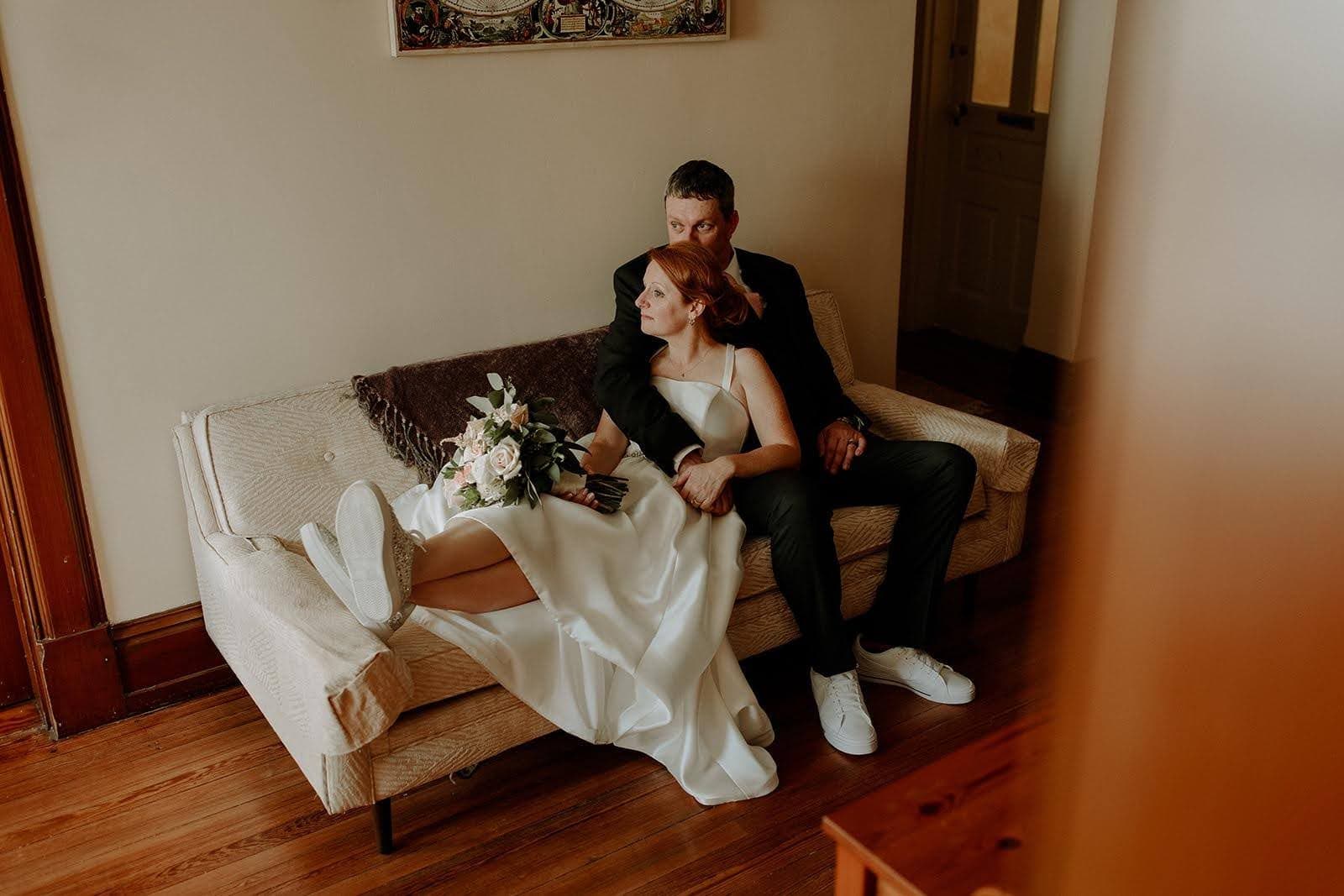 A bride in a simple white dress sits on a couch with her groom, holding a bouquet of flowers.