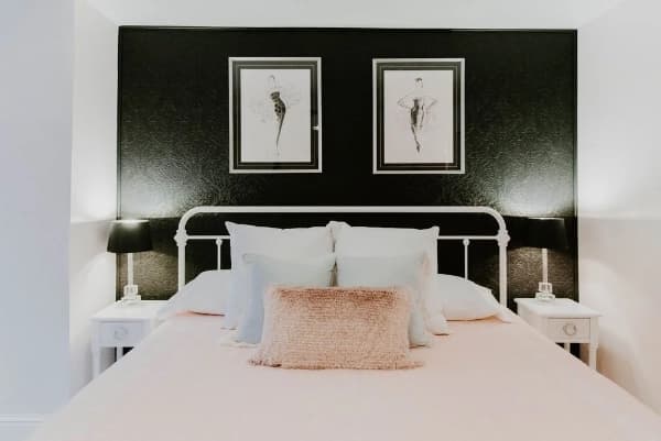 A stylish bedroom featuring a soft pink bed, decorative pillows, and two framed fashion illustrations on a black accent wall.