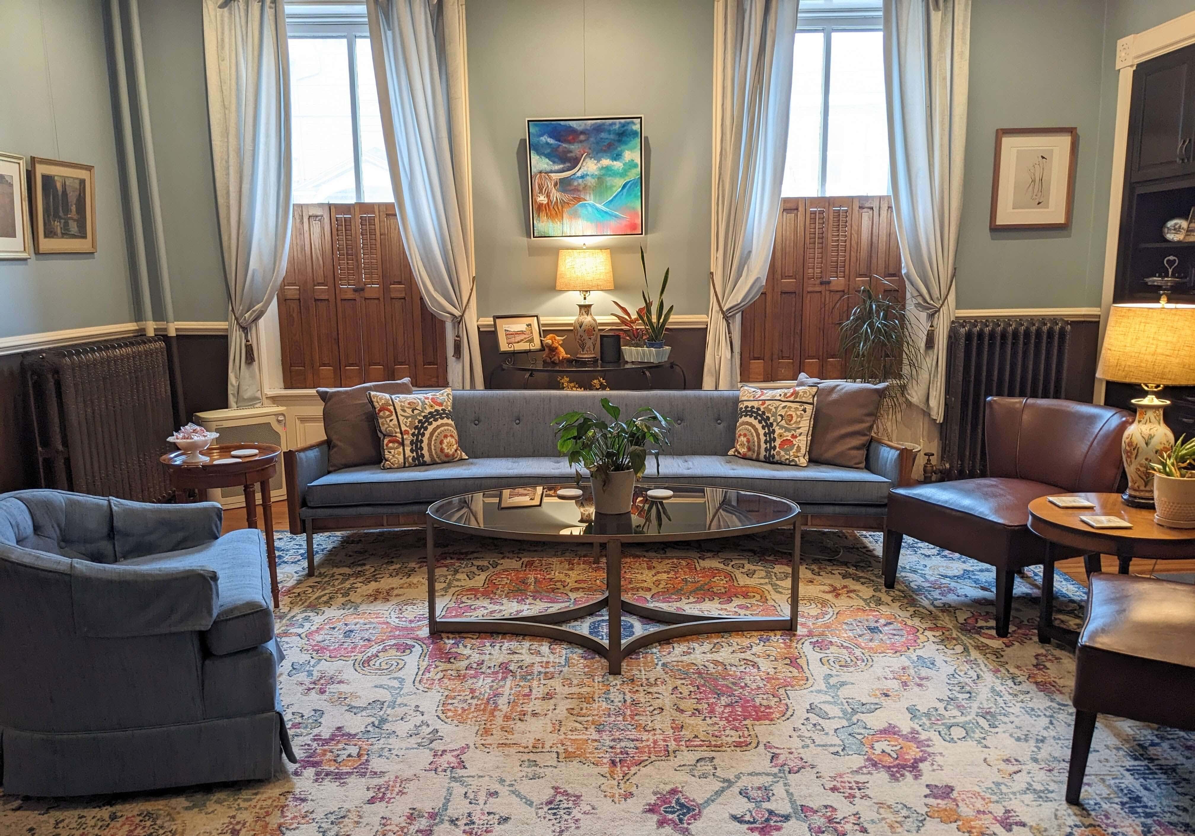 A cozy living room featuring a blue couch, armchairs, a coffee table, and decorative plants, with natural light from large windows.