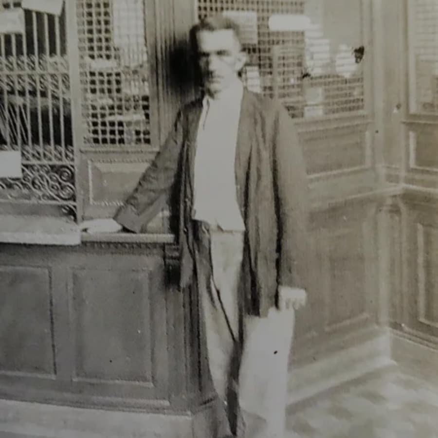 A man in early 20th-century attire stands in a bank, leaning against a counter.