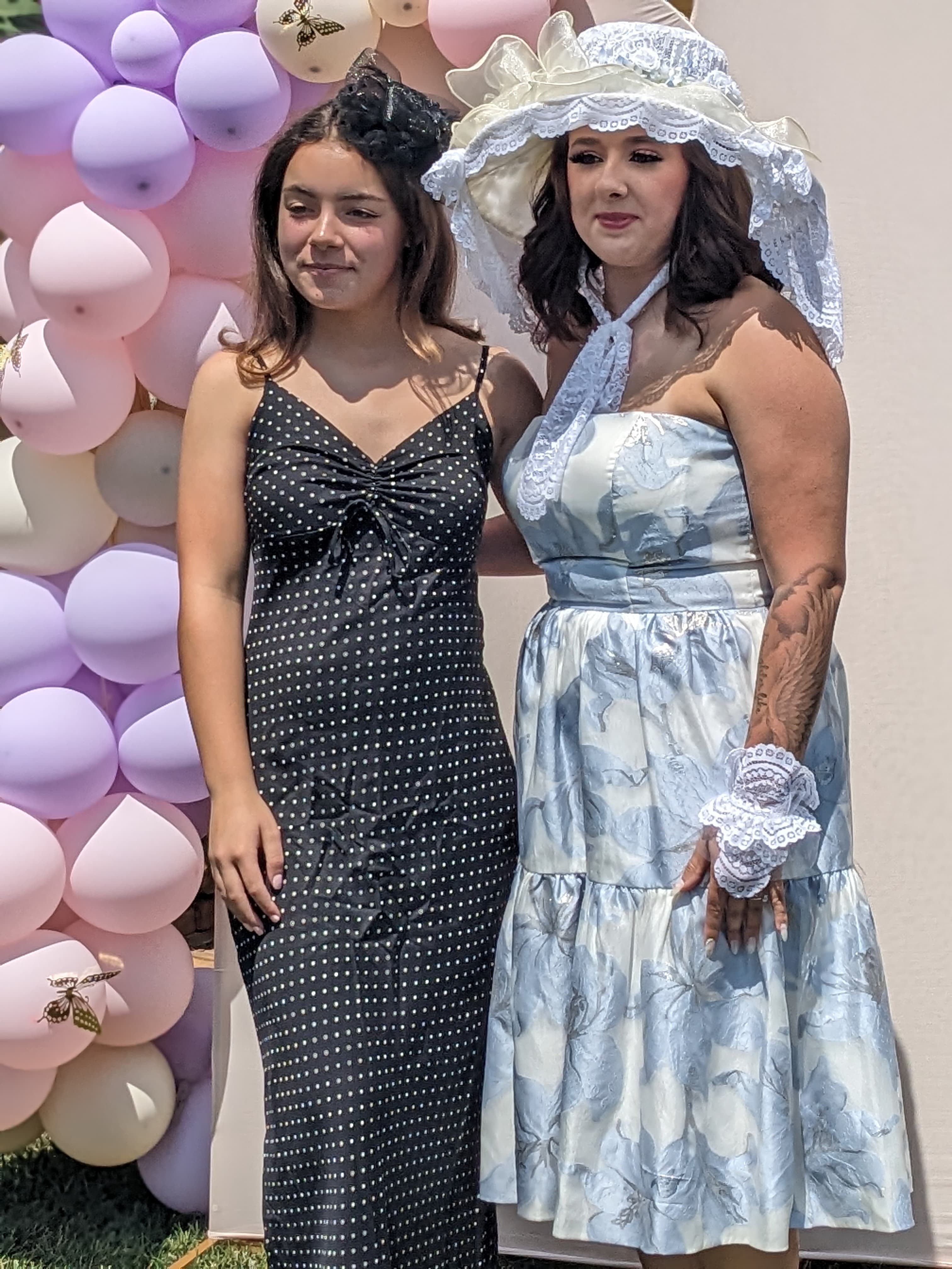 Two women pose for a photo in front of a balloon backdrop, one in a polka dot dress and the other in a floral-patterned dress and large hat.