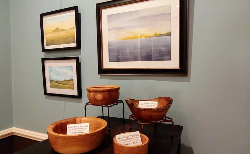 A display of wooden bowls on a table with framed landscape artworks on the wall behind.