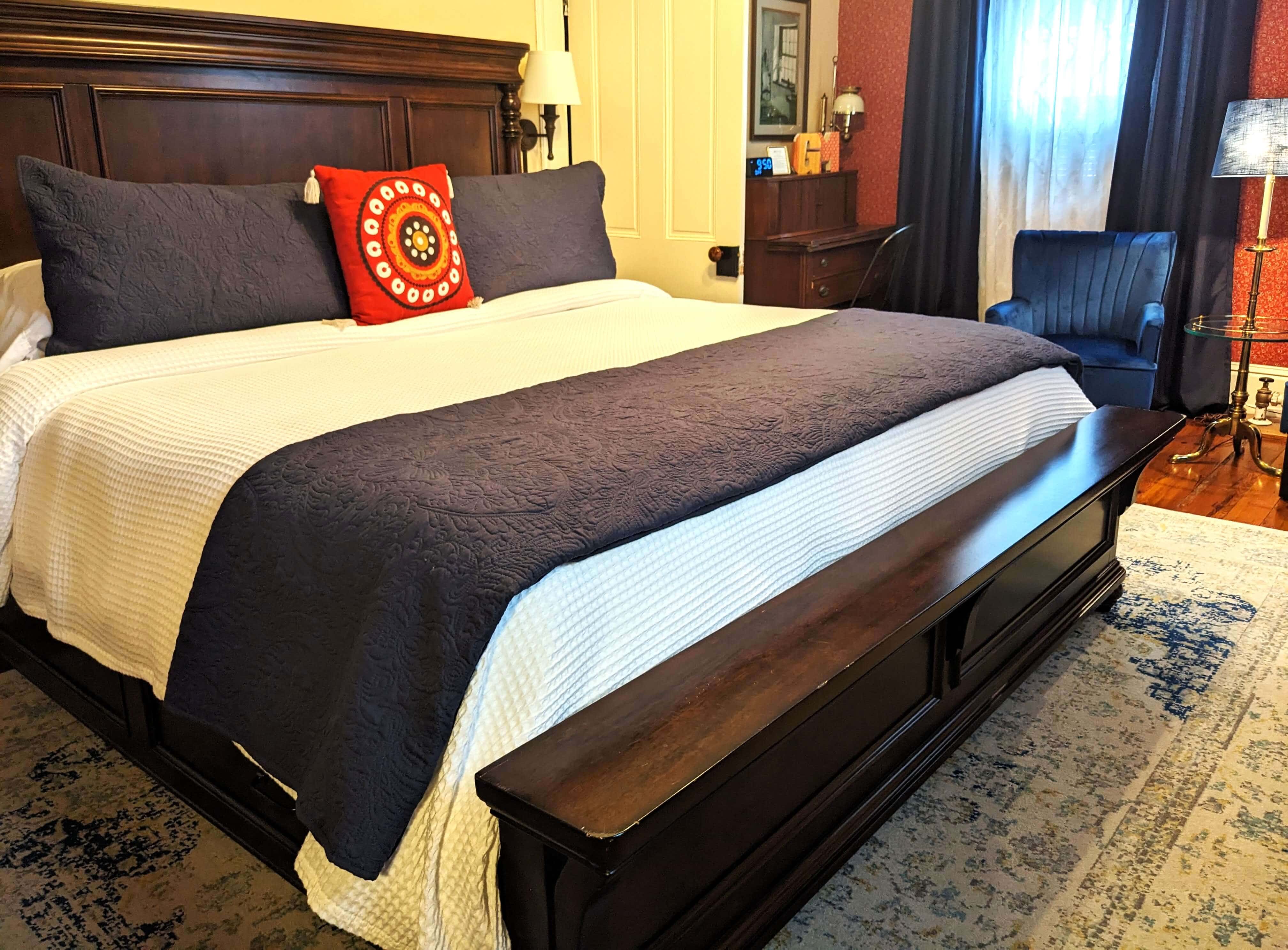 A cozy bedroom featuring a large bed with dark blue bedding and a decorative red pillow, alongside a blue chair and a patterned rug.