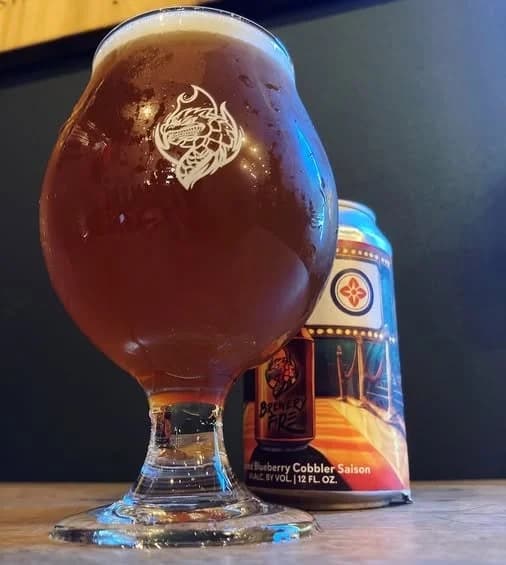 A glass of brown beer sits in front of a colorful can labeled "Blueberry Cobbler Saison." A glass of brown beer sits in front of a colorful can labeled "Blueberry Cobbler Saison."