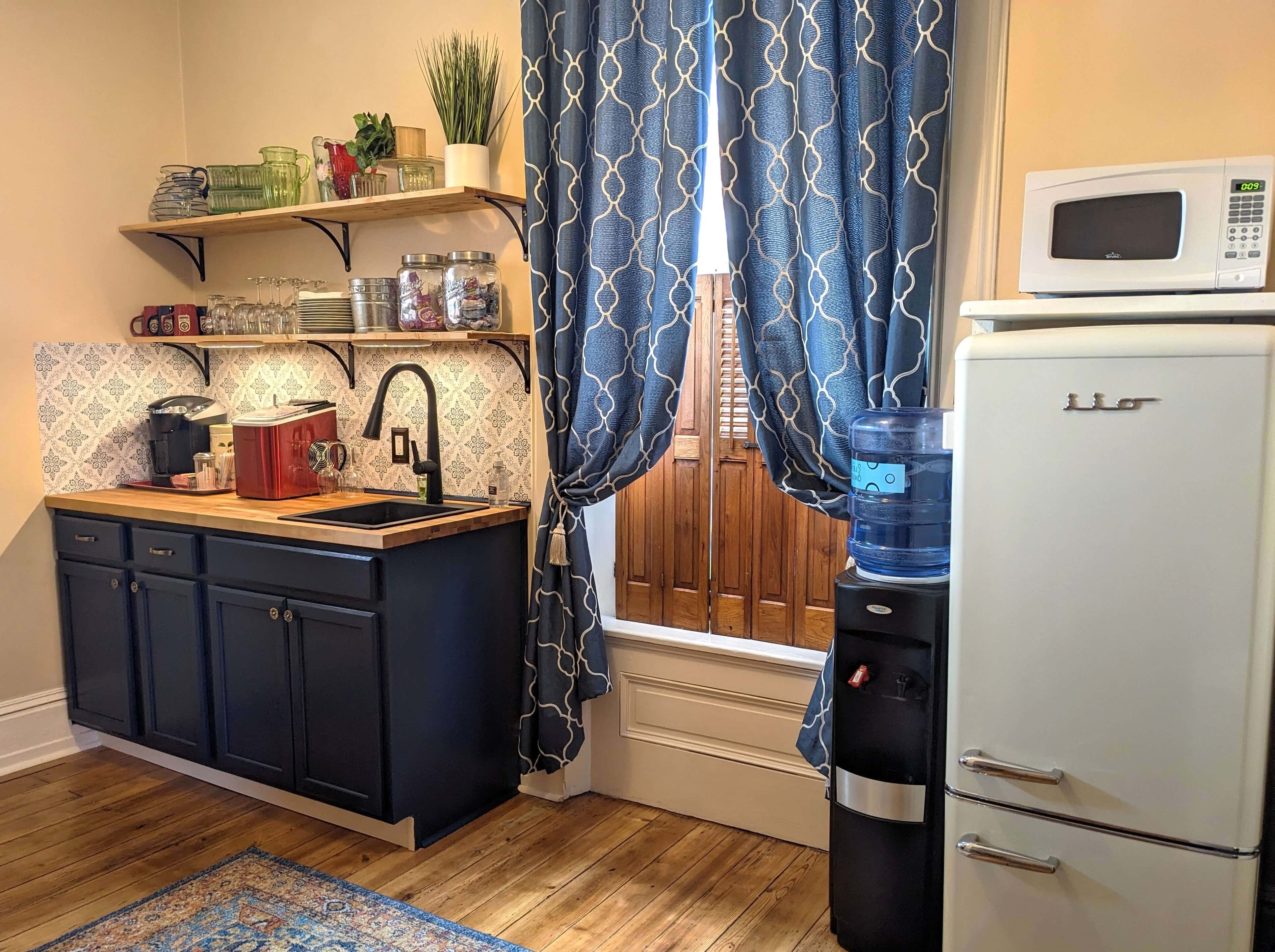 A cozy kitchen featuring dark cabinets, a wooden countertop, and blue patterned curtains with a refrigerator and water cooler.