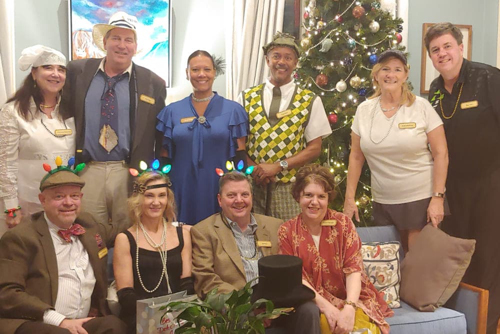 A diverse group of people dressed in festive attire poses together in front of a decorated Christmas tree.