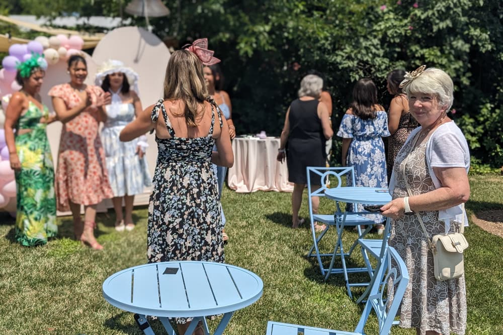 A group of women in dresses socialize at an outdoor gathering, while a woman in a white cardigan and glasses stands near tables.