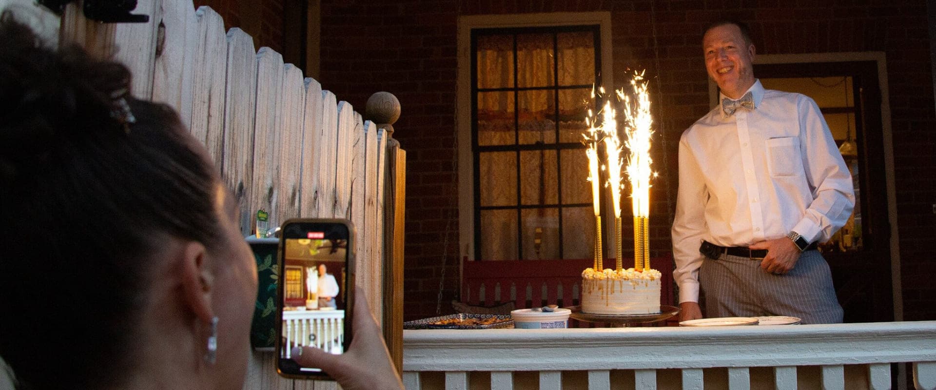 A man in formal attire stands smiling next to a cake with sparklers while a woman takes a photo with her phone.