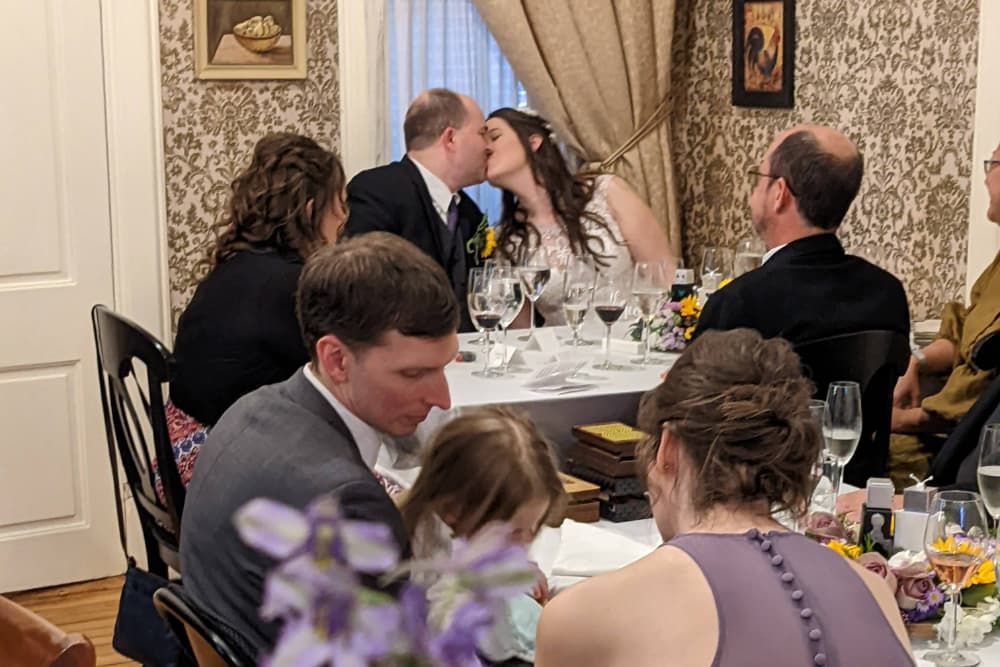 A newlywed couple shares a kiss at their wedding reception, surrounded by guests at a beautifully set table.