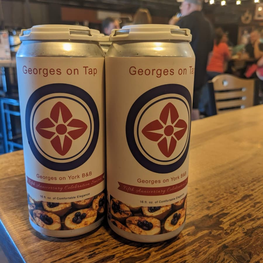 Two cans of beer labeled "Georges on Tap" celebrating the fifth anniversary of "Georges on York B&B," with a wooden table and people in the background.