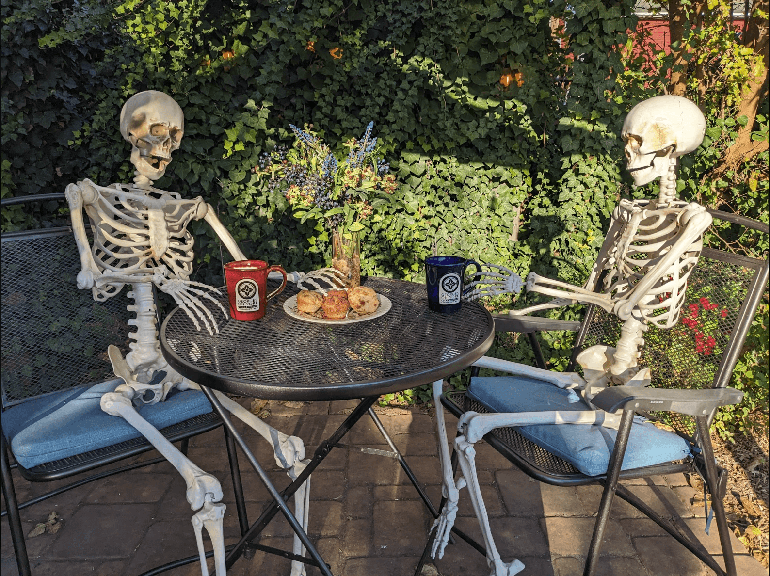 Two skeletons sit at a table with mugs and pastries in a garden setting.